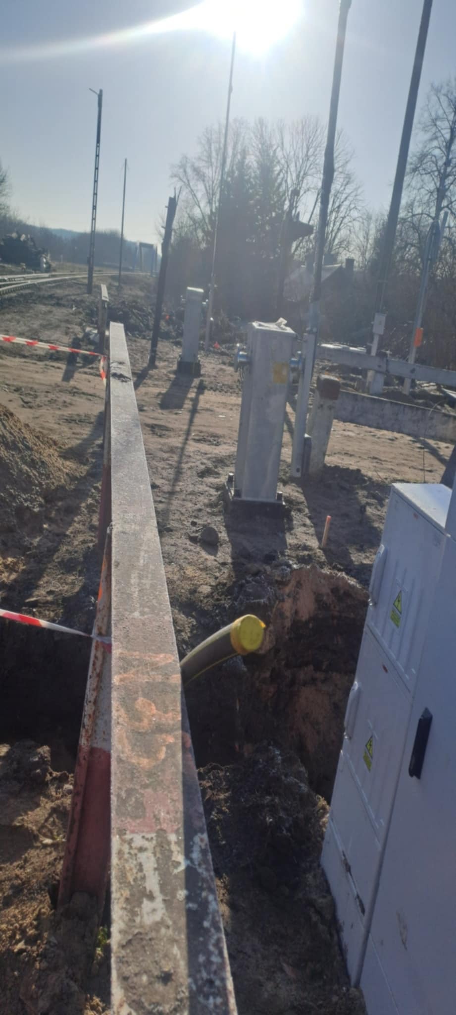 Wykop z rurą w ziemi, obok skrzynka elektryczna i słupy energetyczne w tle. Prace ziemne przy infrastrukturze energetycznej.