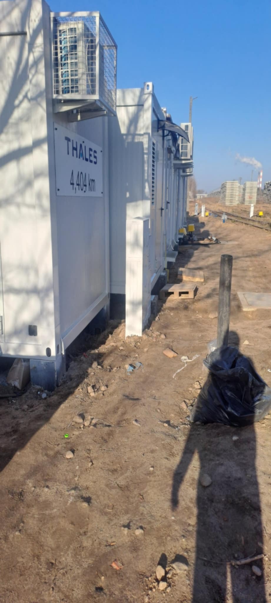 Białe kontenery z klimatyzacją ustawione w rzędzie na placu budowy, widoczne elementy instalacji i cień osoby na ziemi. W tle komin i składowane płyty.