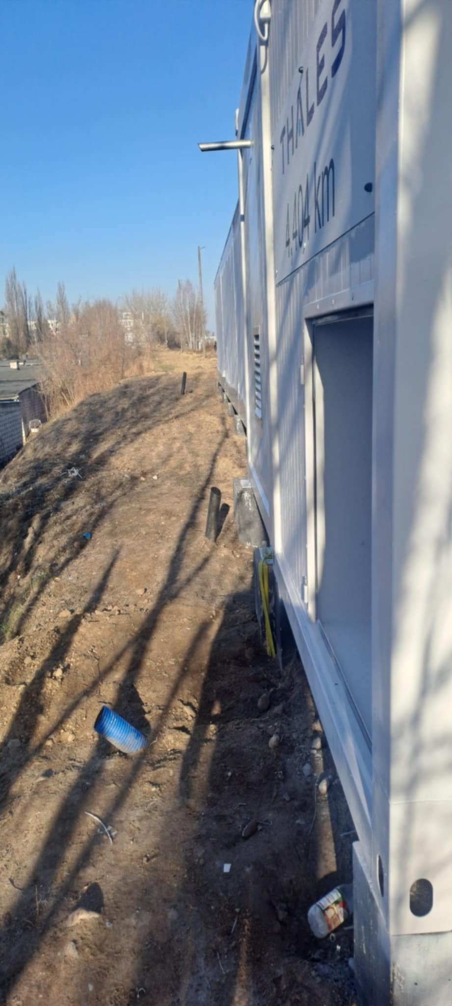 Biały kontener Thales z otwartymi drzwiami, ustawiony na nierównym terenie z widocznymi śladami prac ziemnych i elementami instalacji. Błękitne niebo w tle.