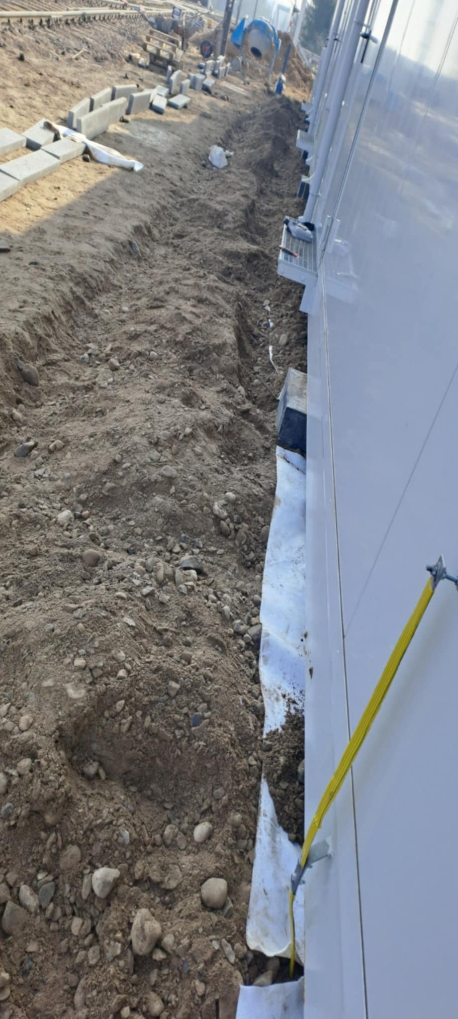 Przygotowanie terenu pod kostkę brukową: wykop, geowłóknina i obrzeża ułożone wzdłuż ściany budynku. Widoczne narzędzia i materiały budowlane w tle.