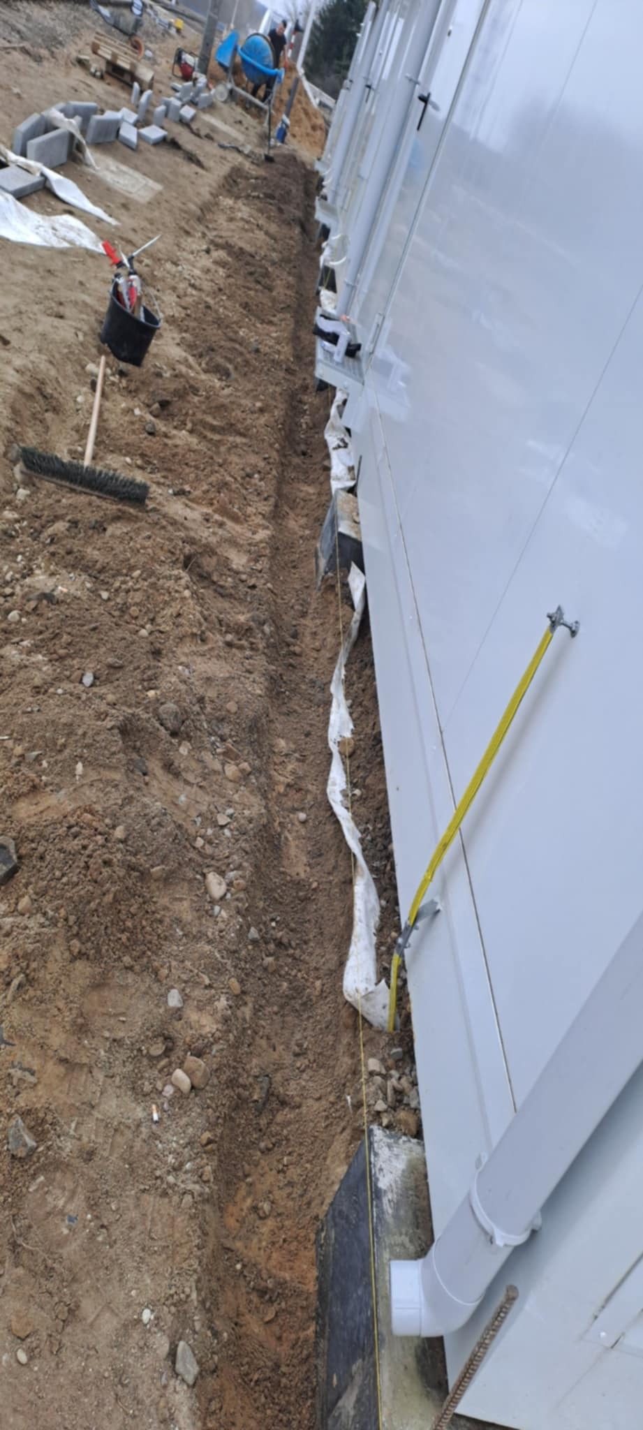 Wykop w ziemi wzdłuż białej ściany budynku, przygotowany pod fundament. Widoczne narzędzia i materiały budowlane, w tle betoniarka i ułożona kostka brukowa.