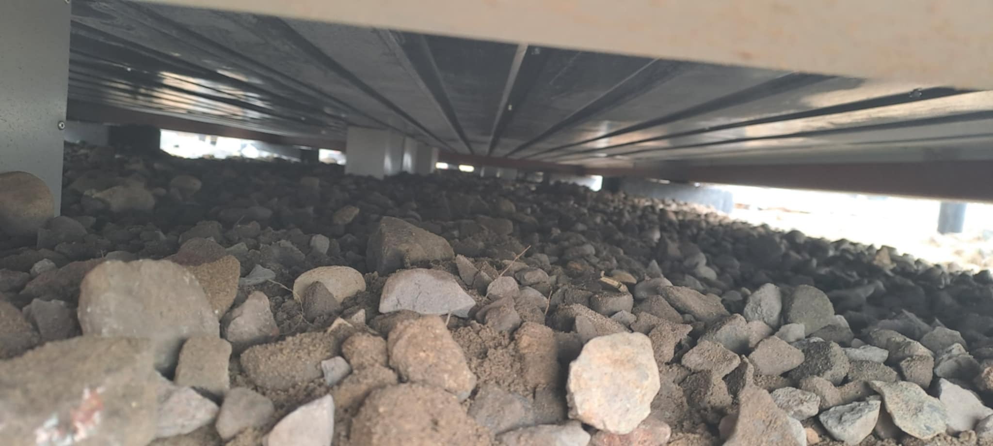 Podsypka z kamieni pod konstrukcją, widoczne elementy wsporcze. Nisko położony kadr, perspektywa od dołu, szaro-brązowa kolorystyka.