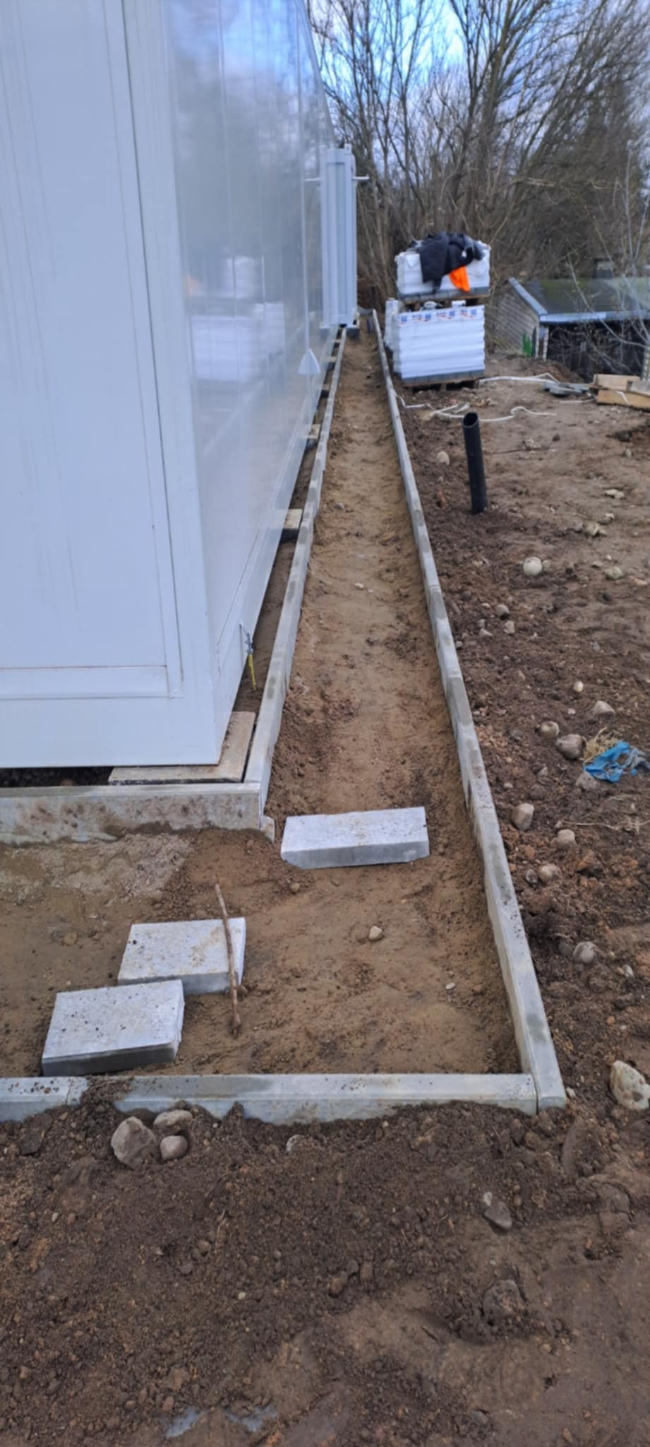 Przygotowanie podłoża pod ścieżkę z kostki brukowej. Widoczne obrzeża i częściowo ułożone płyty. W tle budynek kontenerowy i materiały budowlane.