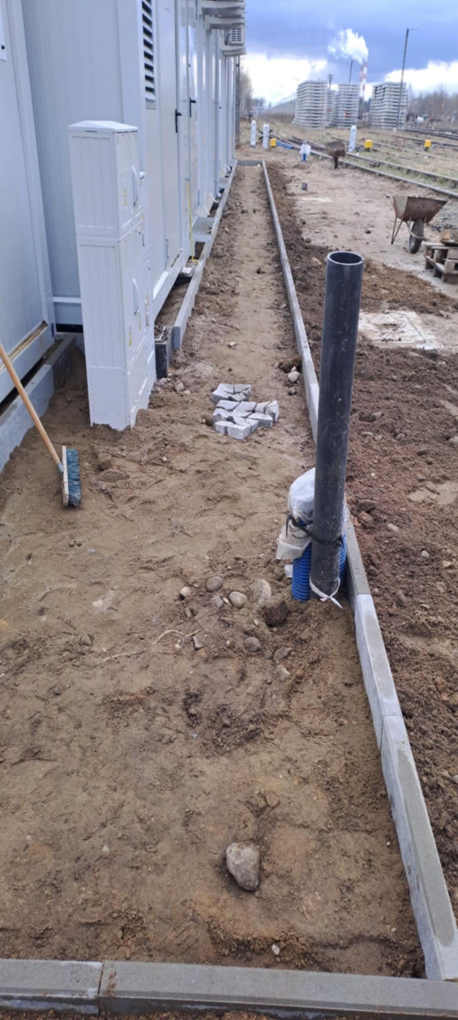 Przygotowanie terenu pod ścieżkę z kostki brukowej: ułożone obrzeża, rozsypana ziemia, widoczne kostki, rura kanalizacyjna i szczotka. W tle budynki i teren budowy.