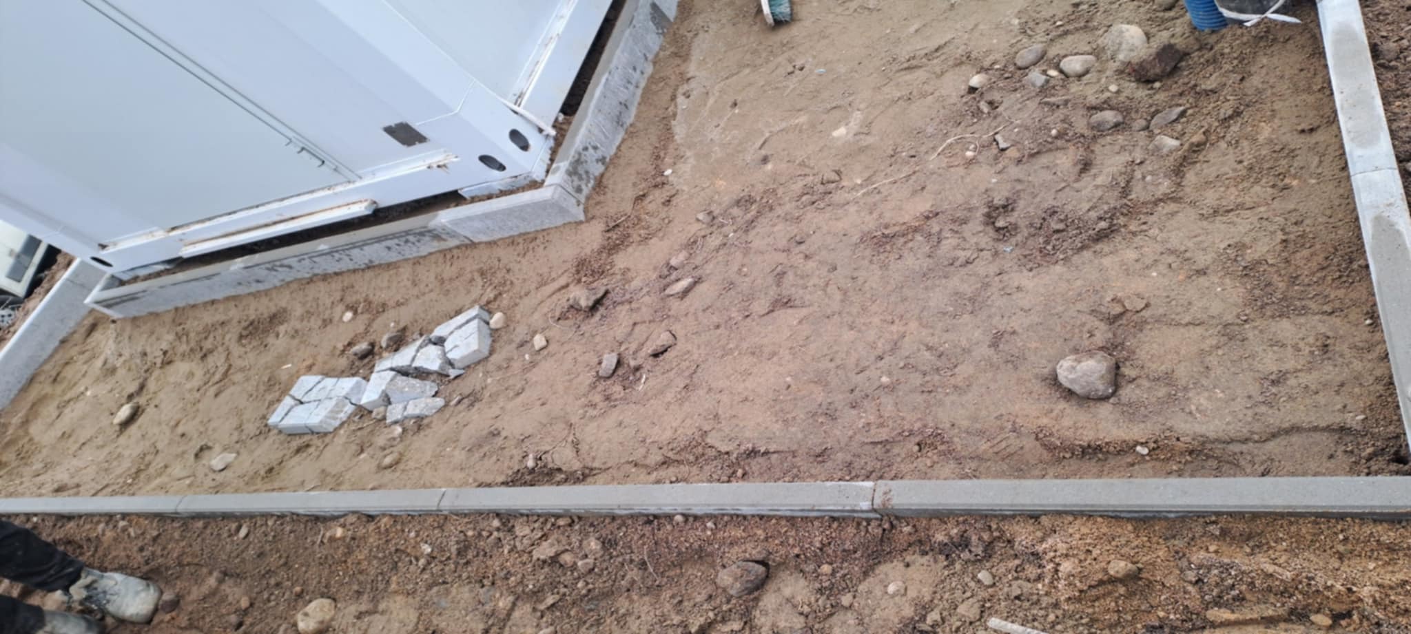 Etap układania kostki granitowej: obrzeża ułożone, podłoże przygotowane, kostka częściowo ułożona przy budynku. Widoczne narzędzia i materiały budowlane.