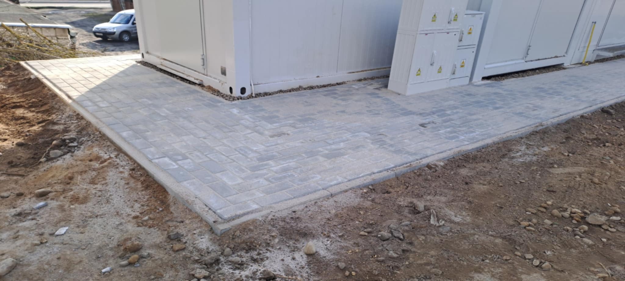 Ułożona szara kostka brukowa w jodełkę przy białym kontenerze, widoczne skrzynki elektryczne. Krawężnik oddziela kostkę od ziemi. W tle zaparkowany samochód.