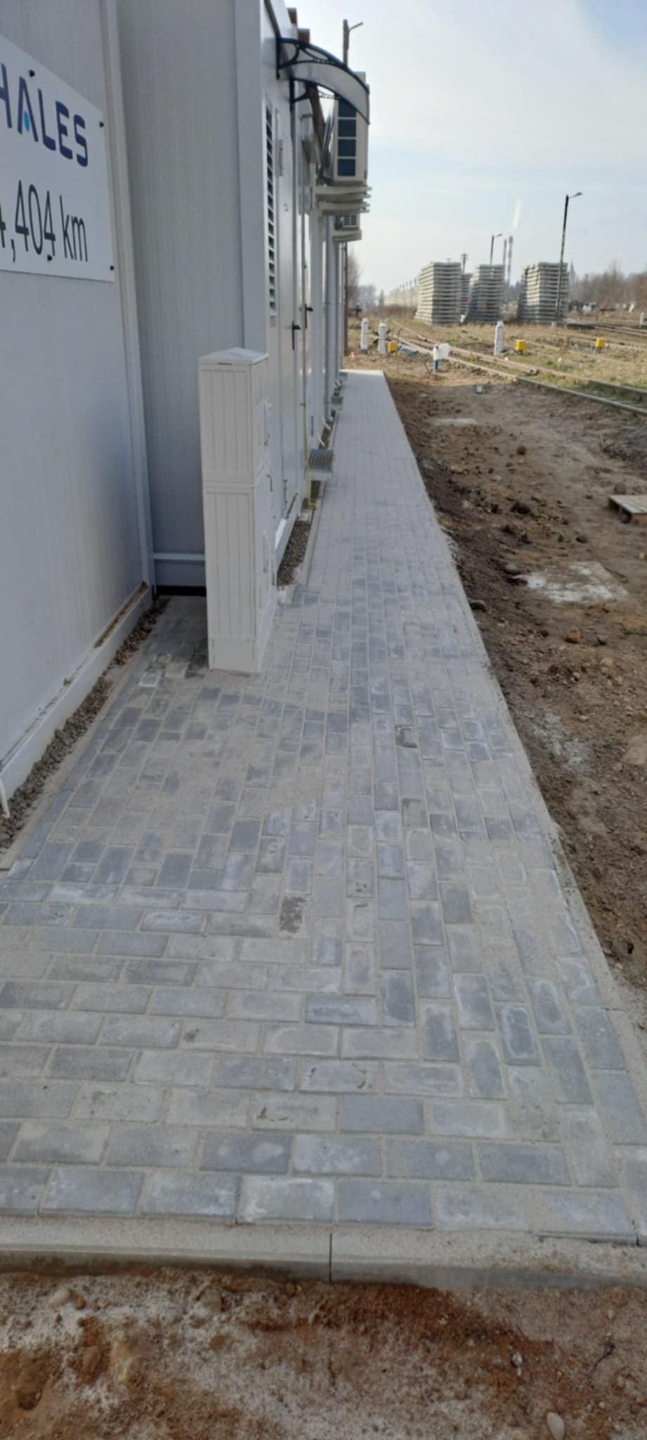 Ścieżka z szarej kostki brukowej przy budynku kontenerowym w Białystoku. Widoczne elementy infrastruktury i teren przygotowany pod dalsze prace.