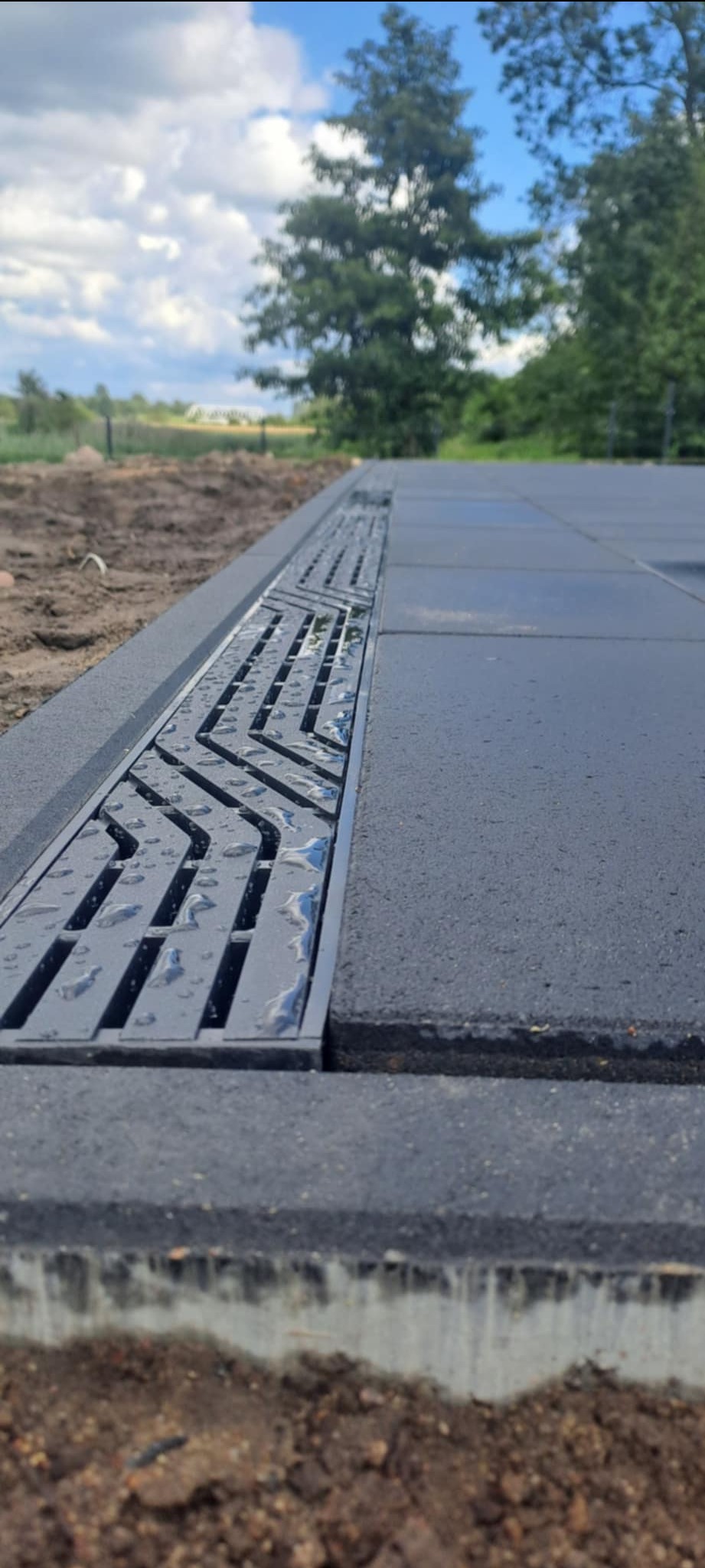 Ułożona, szara kostka brukowa z odpływem liniowym w kształcie zygzaka, na którym widoczne są krople wody. W tle ziemia i zieleń.