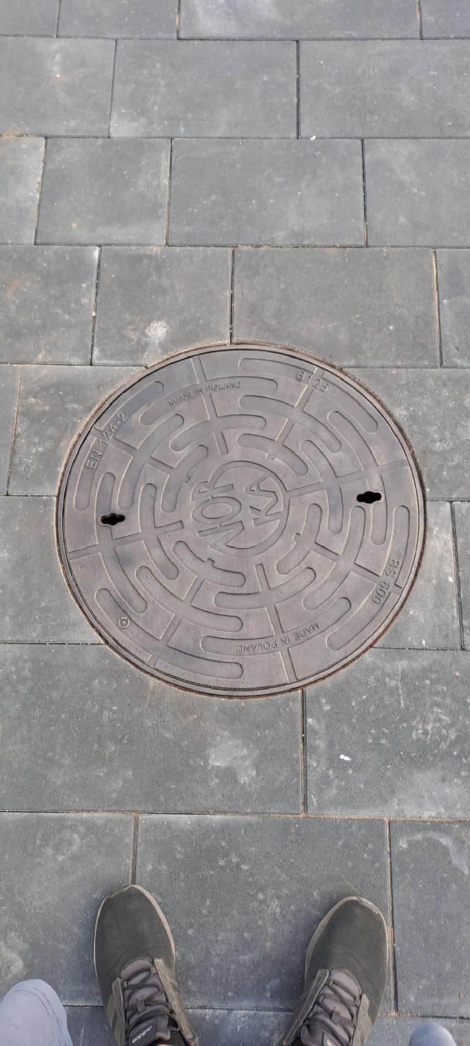 Szara kostka brukowa z okrągłą studzienką kanalizacyjną z napisem 'Made in Poland' i butami w dolnej części kadru.