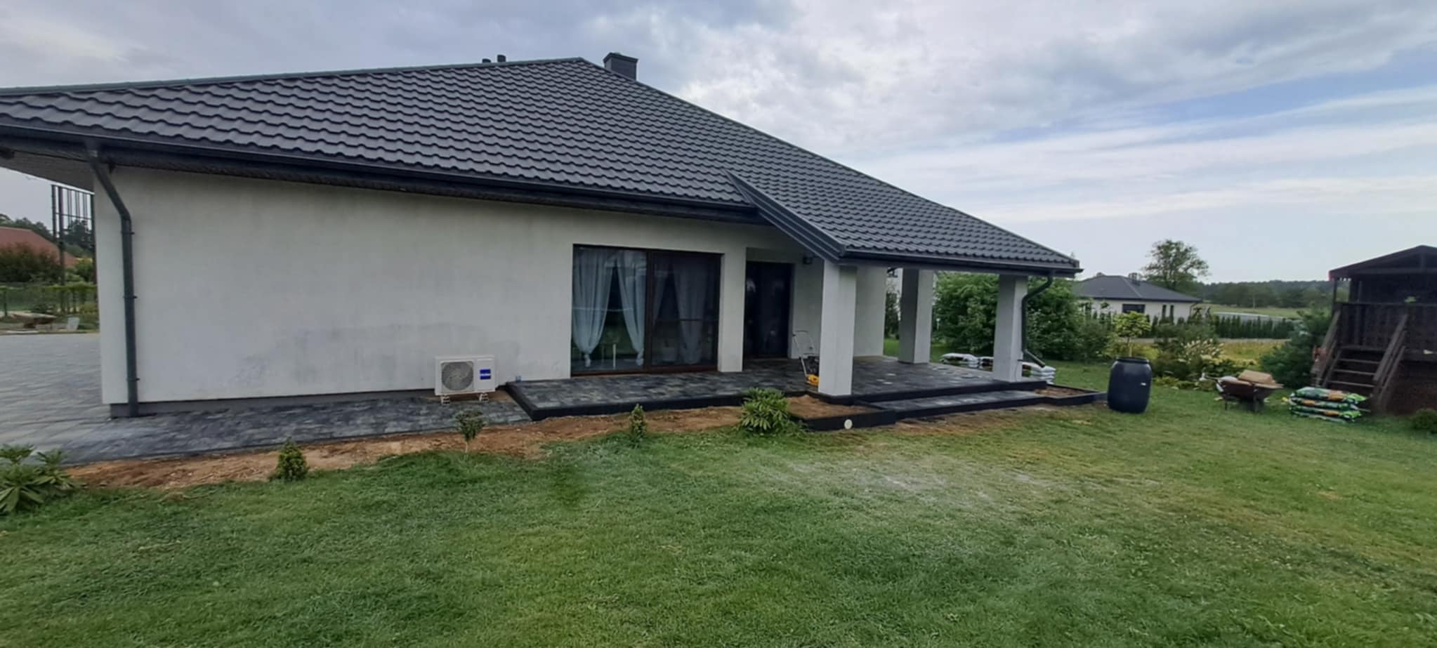 Nowoczesny dom z ciemną kostką brukową na tarasie i wokół budynku. Widoczne elementy wykończeniowe i zagospodarowanie terenu. Ujęcie z poziomu trawnika.