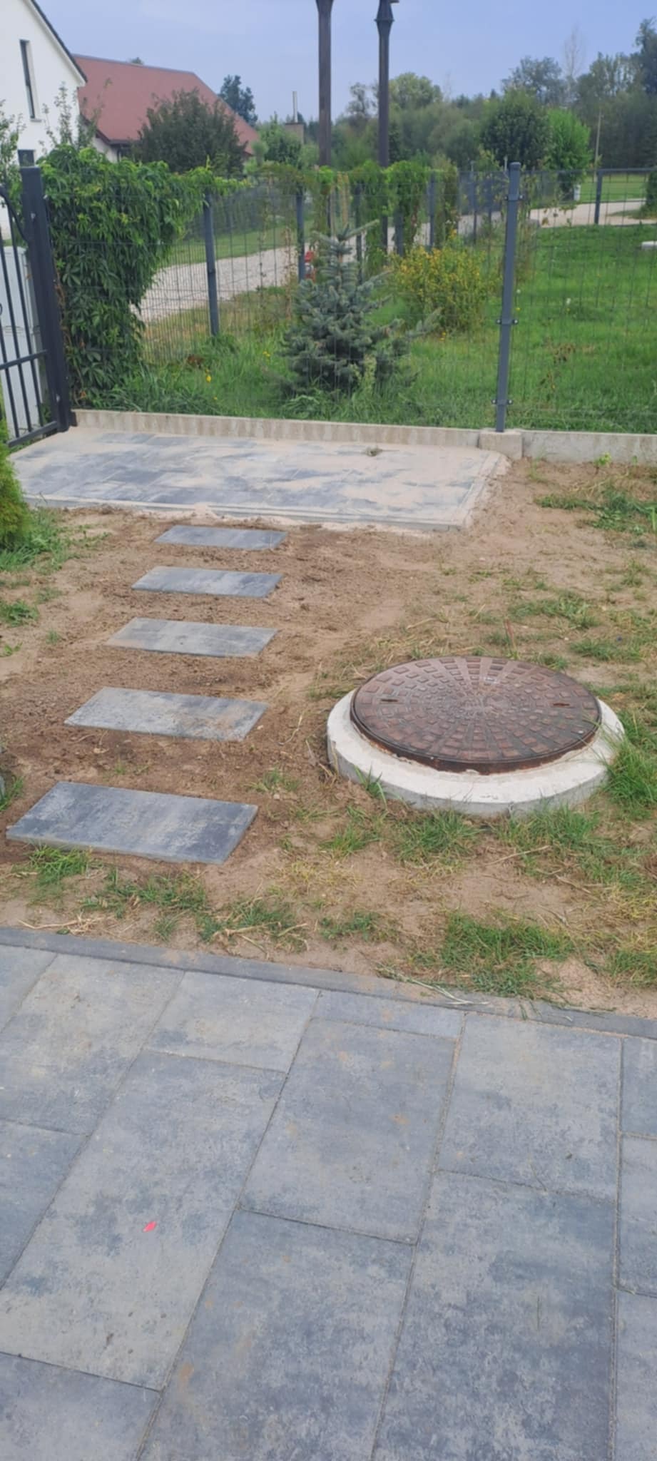 Ścieżka z płyt kamiennych prowadząca do studzienki kanalizacyjnej na trawniku, obok ułożona kostka brukowa. Widok na ogród i ogrodzenie.