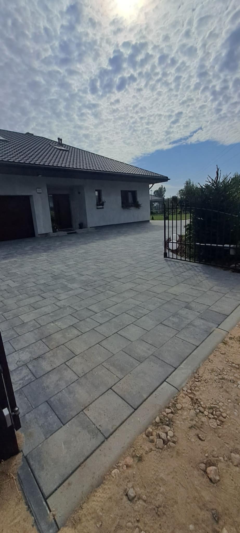 Ułożona szara kostka brukowa na podjeździe przed domem jednorodzinnym z bramą w tle, widok z perspektywy lekko z boku, w słoneczny dzień.