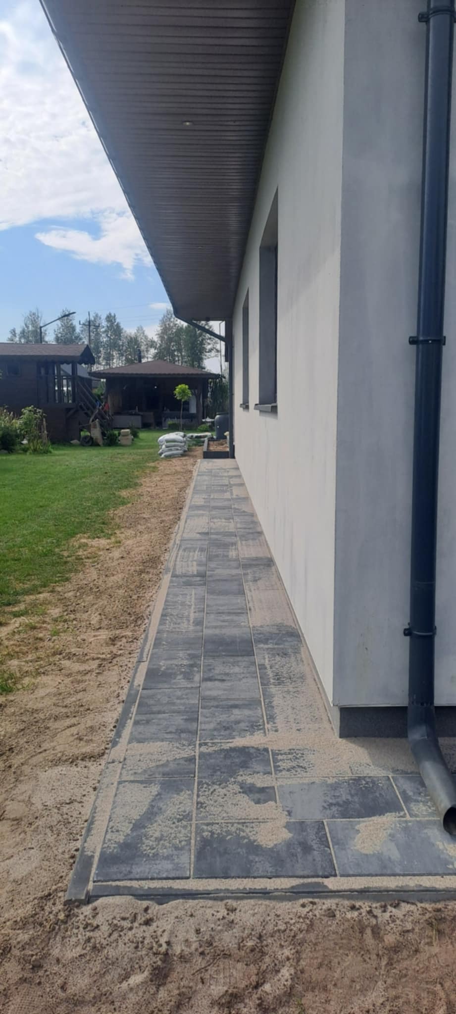 Ułożona, szara kostka brukowa tworząca ścieżkę wzdłuż jasnego budynku z rynną. W tle widoczny trawnik, drzewa i elementy architektury ogrodowej.