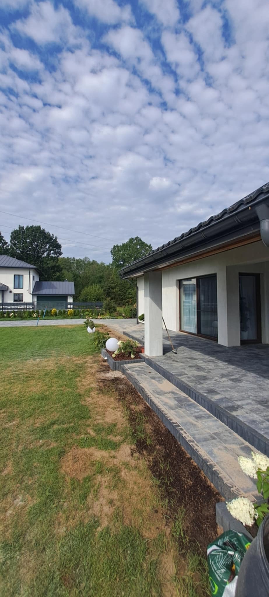 Nowoczesny dom z tarasem wyłożonym szarą kostką brukową. Widoczne schody z kostki, trawnik i elementy dekoracyjne ogrodu. Błękitne niebo z obłokami w tle.