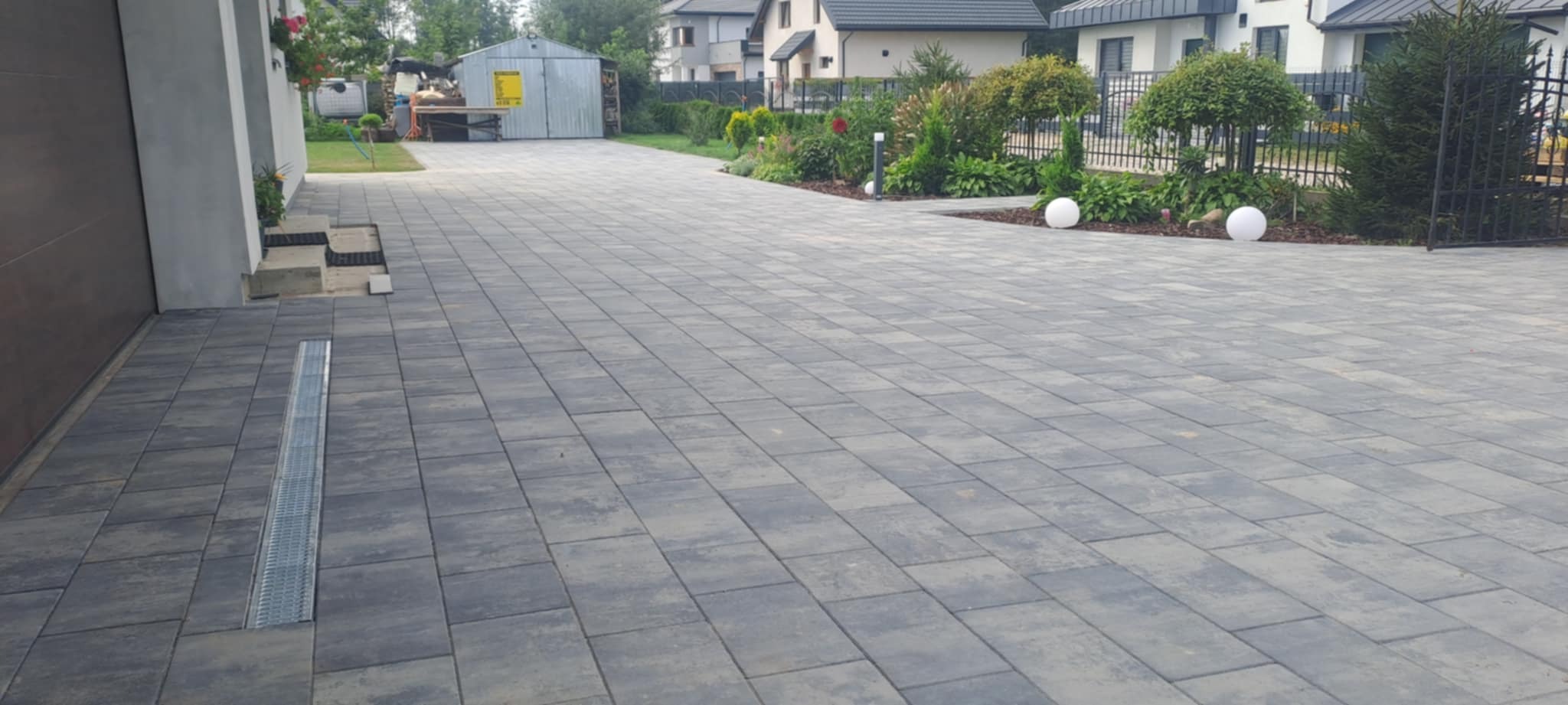 Ułożona, szara kostka brukowa na podjeździe z odpływem liniowym, prowadząca do garażu i ogrodu z dekoracyjnymi kulami. Widok na domy w tle.
