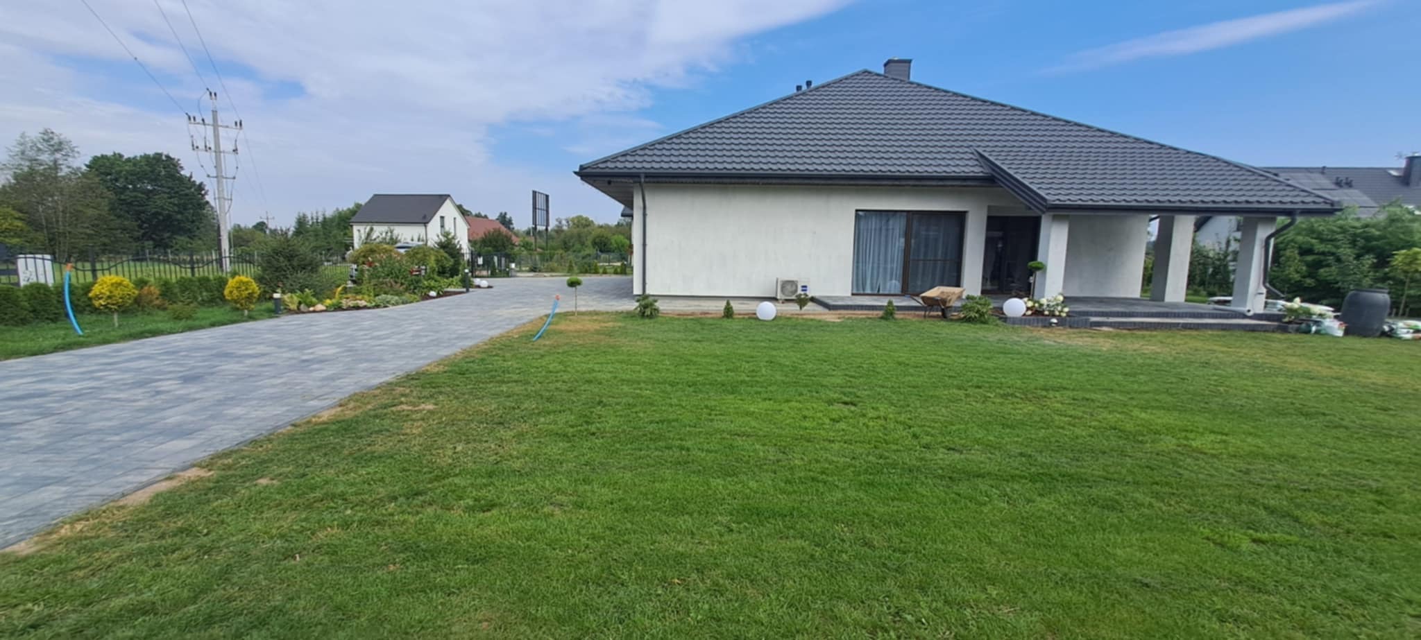 Nowoczesny dom jednorodzinny z szarą kostką brukową na podjeździe i zadbanym trawnikiem. Widoczne elementy dekoracyjne ogrodu i oświetlenie.