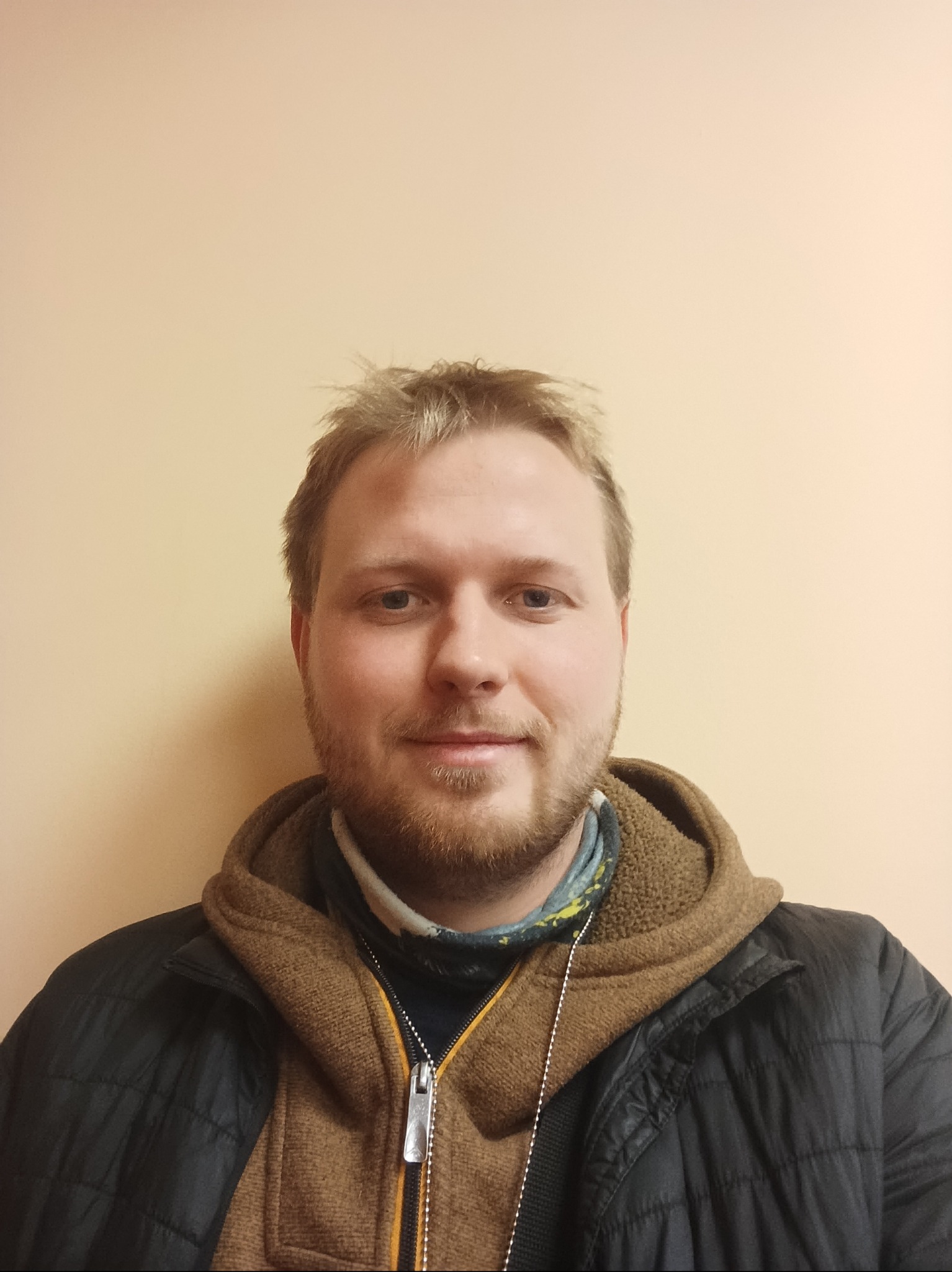 Portret mężczyzny z lekkim zarostem, ubranego w kurtkę i bluzę z kapturem, na jednolitym, jasnym tle. Widoczne detale ubrania i delikatny uśmiech.