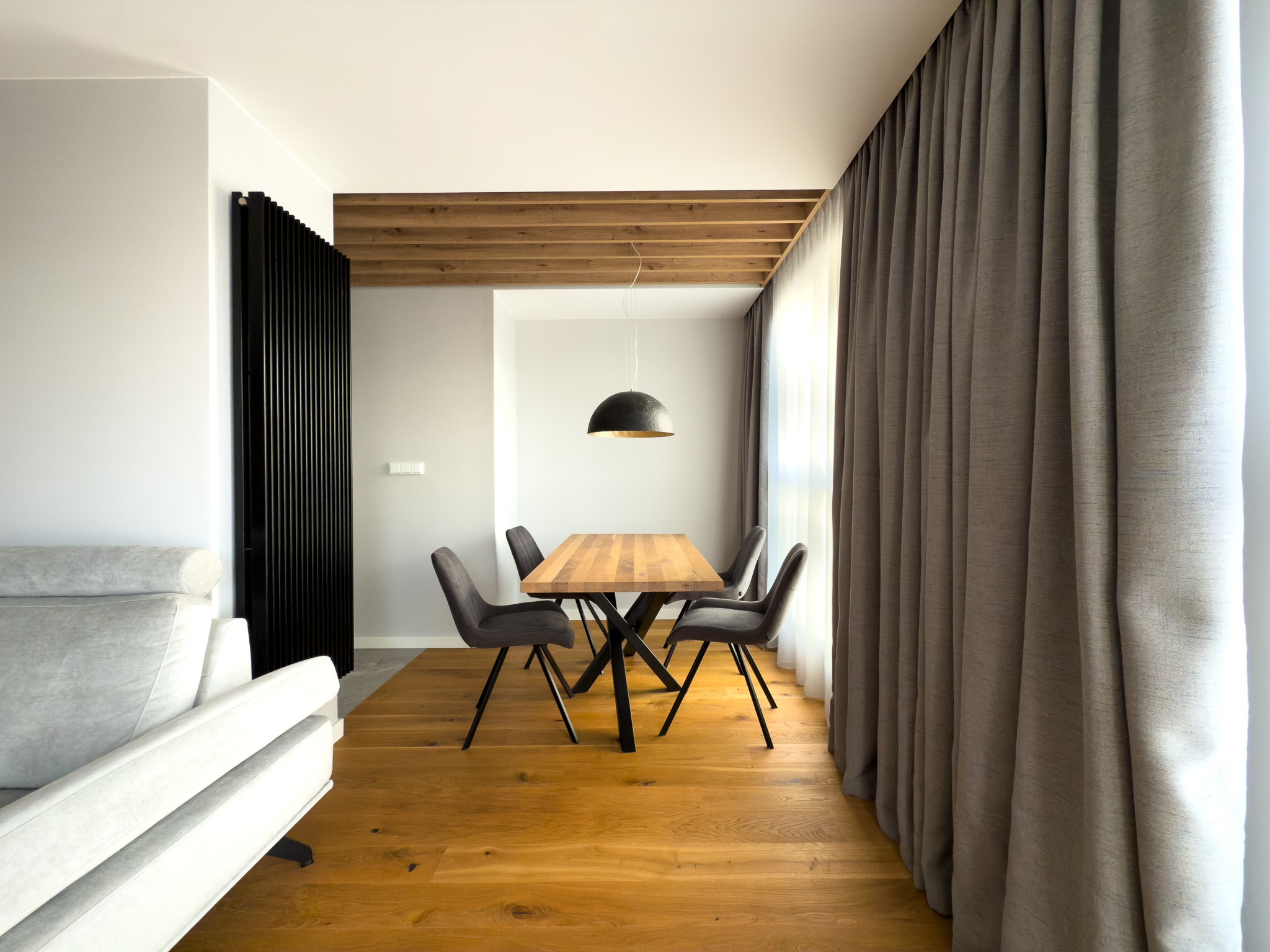 Jasne wnętrze z drewnianą podłogą i stołem z krzesłami. Czarna lampa wisząca nad stołem, szare zasłony i sofa. Drewniany sufit i czarny, dekoracyjny panel.
