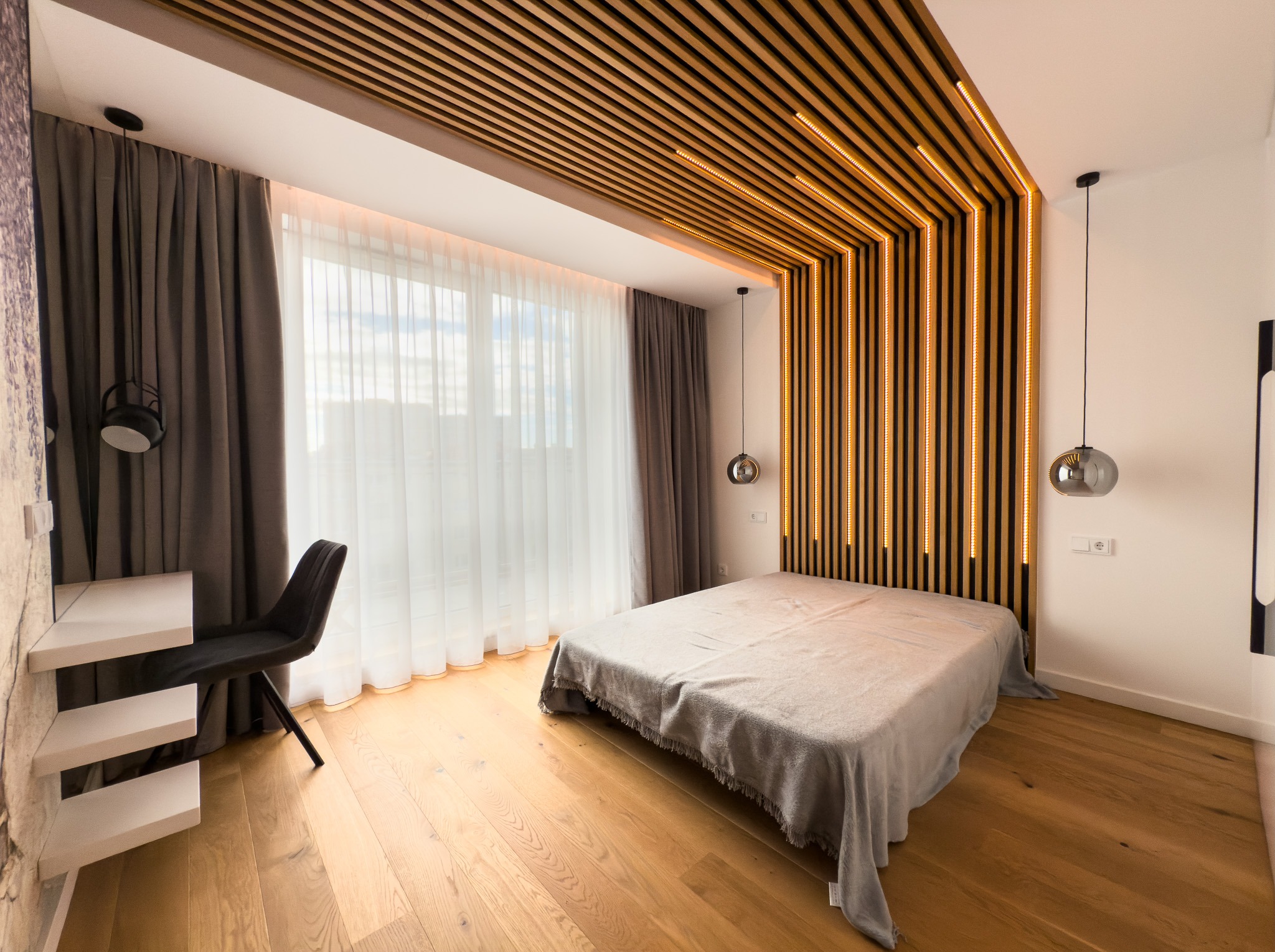 Nowoczesna sypialnia z drewnianym sufitem i oświetleniem LED, biurko z krzesłem przy oknie z delikatnymi zasłonami, parkiet na podłodze i minimalistyczne lampy wiszące.