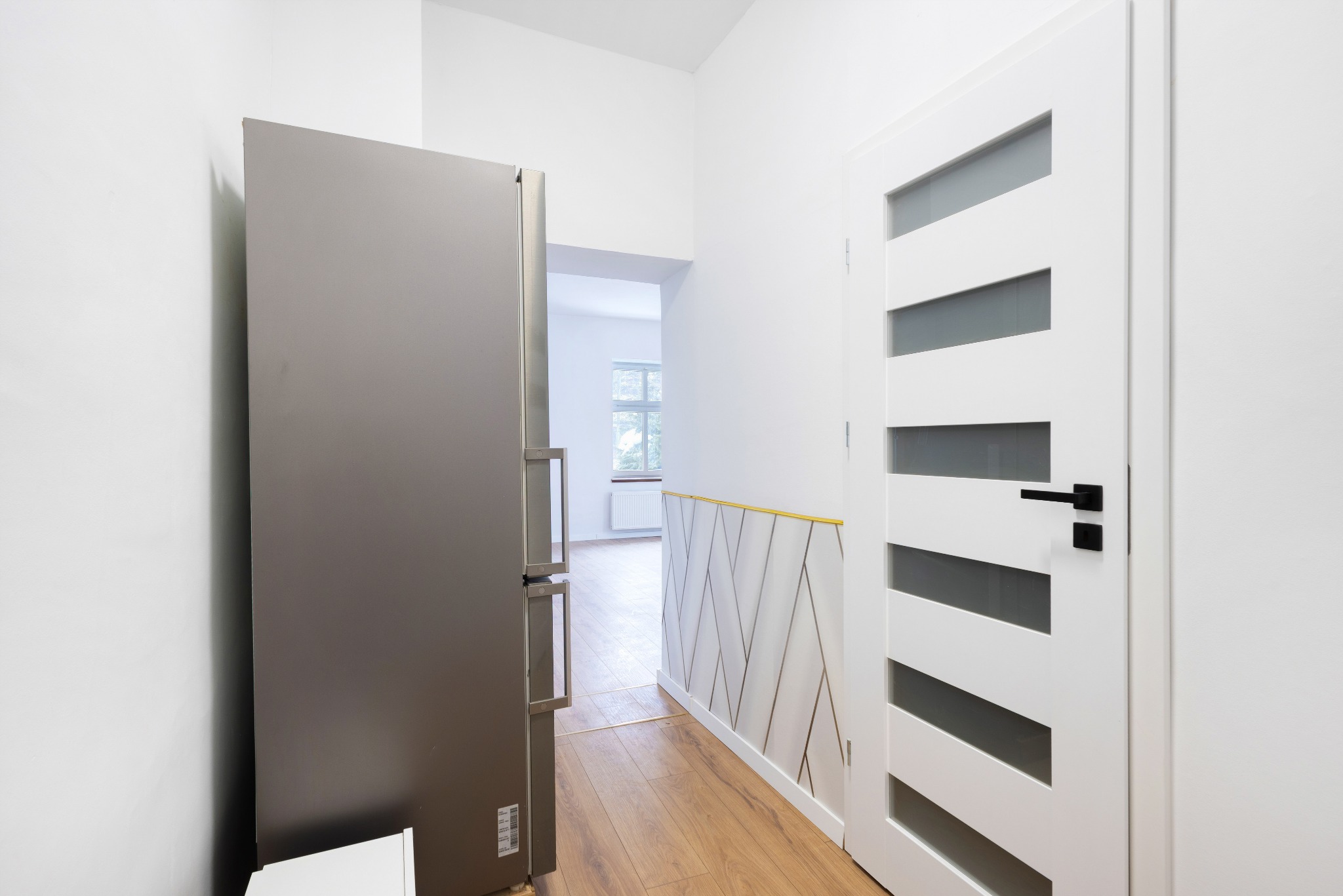 Nowoczesny przedpokój z drewnianą podłogą, białą ścianą z dekoracyjnym panelem i szarą lodówką. Widok na jasne pomieszczenie w głębi.