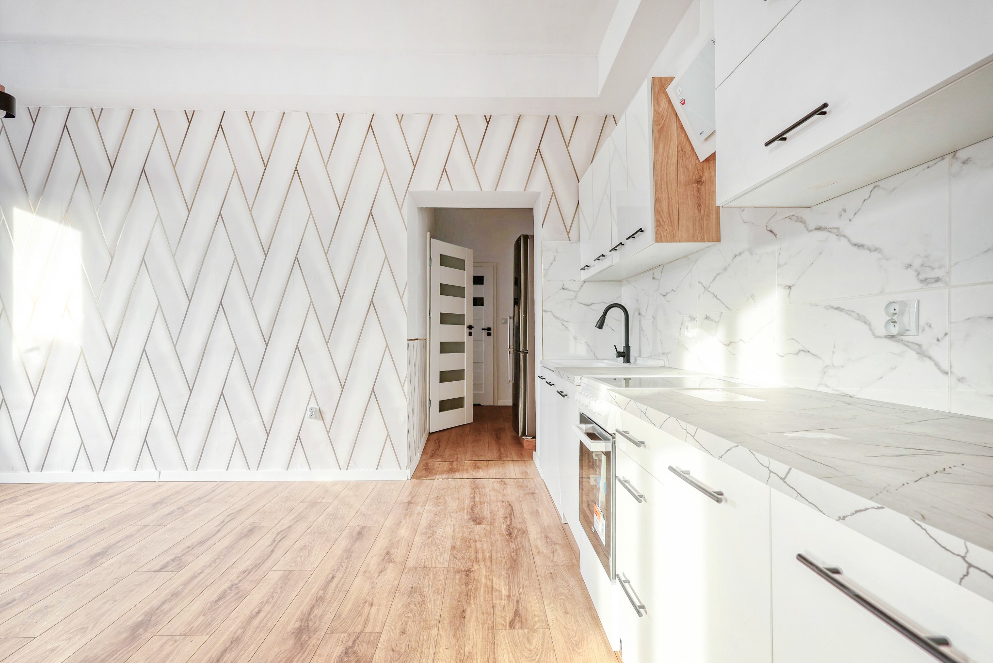 Jasna kuchnia z białymi szafkami i blatem imitującym marmur, czarna armatura, drewniana podłoga i ściana z geometrycznym wzorem. Widok na otwarte drzwi.