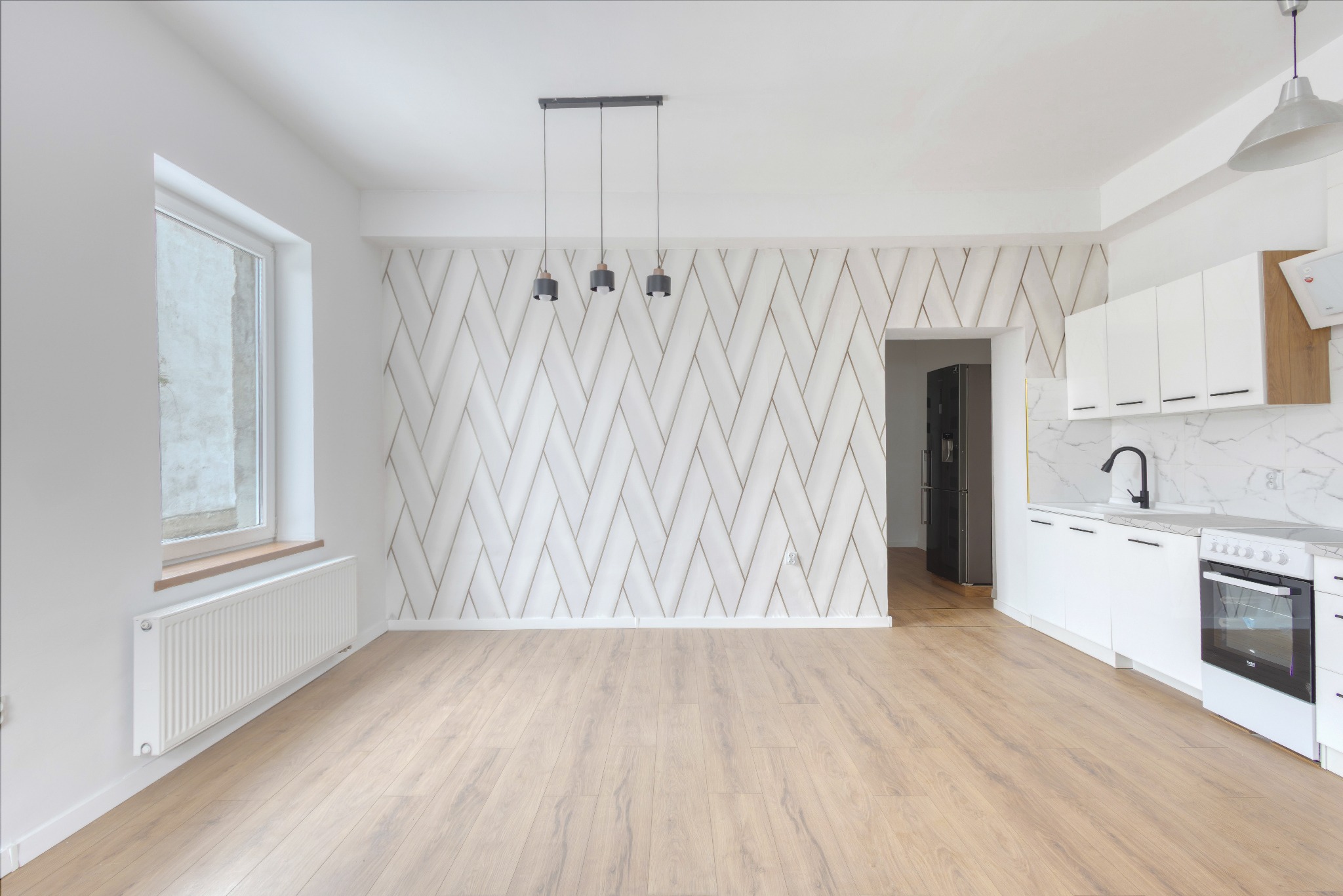 Jasne wnętrze: parkiet, białe ściany z geometrycznym wzorem, okno z grzejnikiem, biała zabudowa kuchenna z czarną armaturą i sprzętami. Minimalistyczny design.
