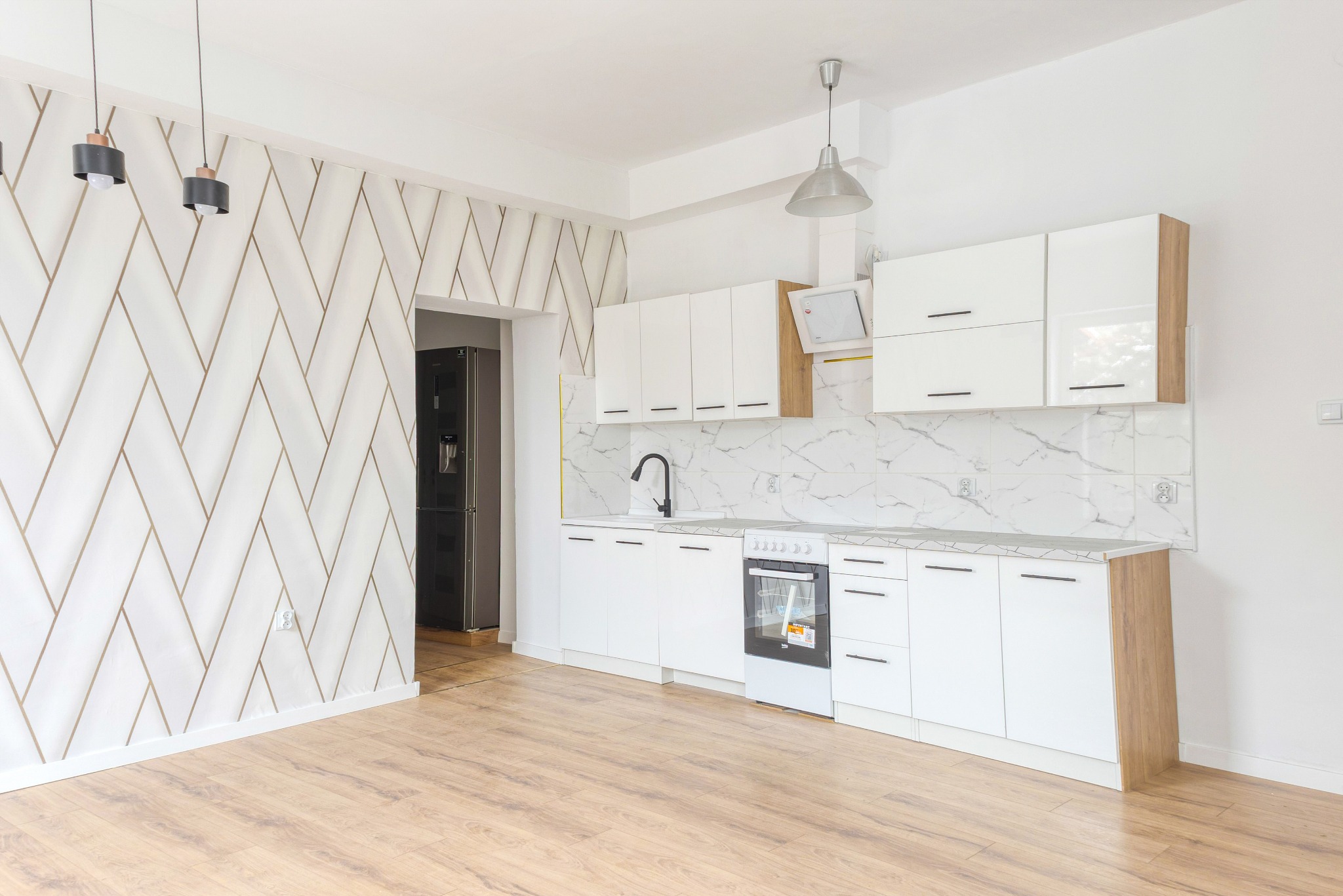 Jasna kuchnia z białymi szafkami, marmurowym blatem i wzorzystą ścianą. Nowoczesne oświetlenie i drewniana podłoga dodają elegancji. Widok na czarną lodówkę w tle.