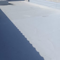 Seweryn Wojciechowski - Szary, płaski dach pokryty membraną, z widocznymi łączeniami i cieniem rzucanym przez krawędź dachu. Minimalistyczny kadr, skupiający się na fakturze i geometrii pokrycia.