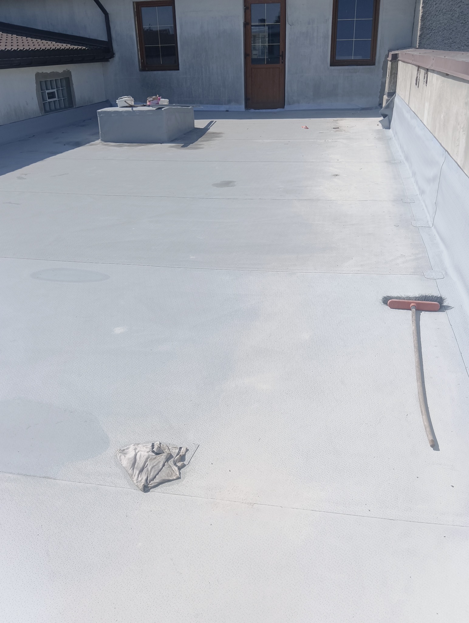 Jasnoszary dach płaski po renowacji z widocznymi narzędziami: szczotką i szmatką. W tle fragment budynku z oknami i drzwiami.