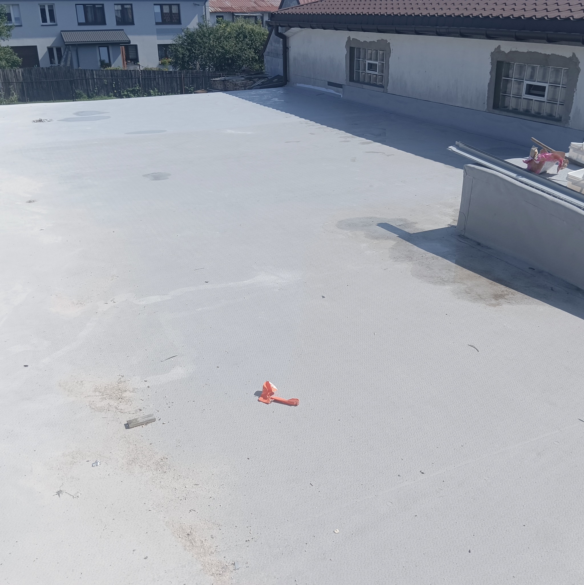 Szary, płaski dach po renowacji z widocznymi śladami prac, narzędziami i elementami wykończeniowymi. W tle budynek z oknami i fragment dachu sąsiedniego domu.