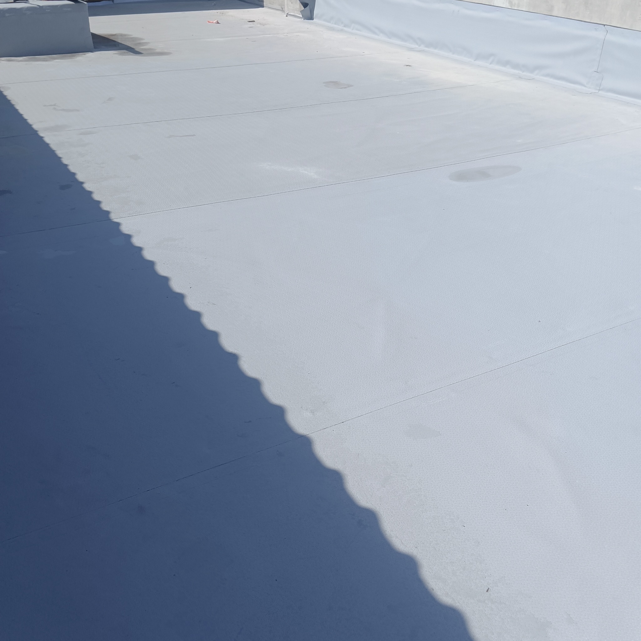 Szary, płaski dach pokryty membraną, z widocznymi łączeniami i cieniem rzucanym przez krawędź dachu. Minimalistyczny kadr, skupiający się na fakturze i geometrii pokrycia.