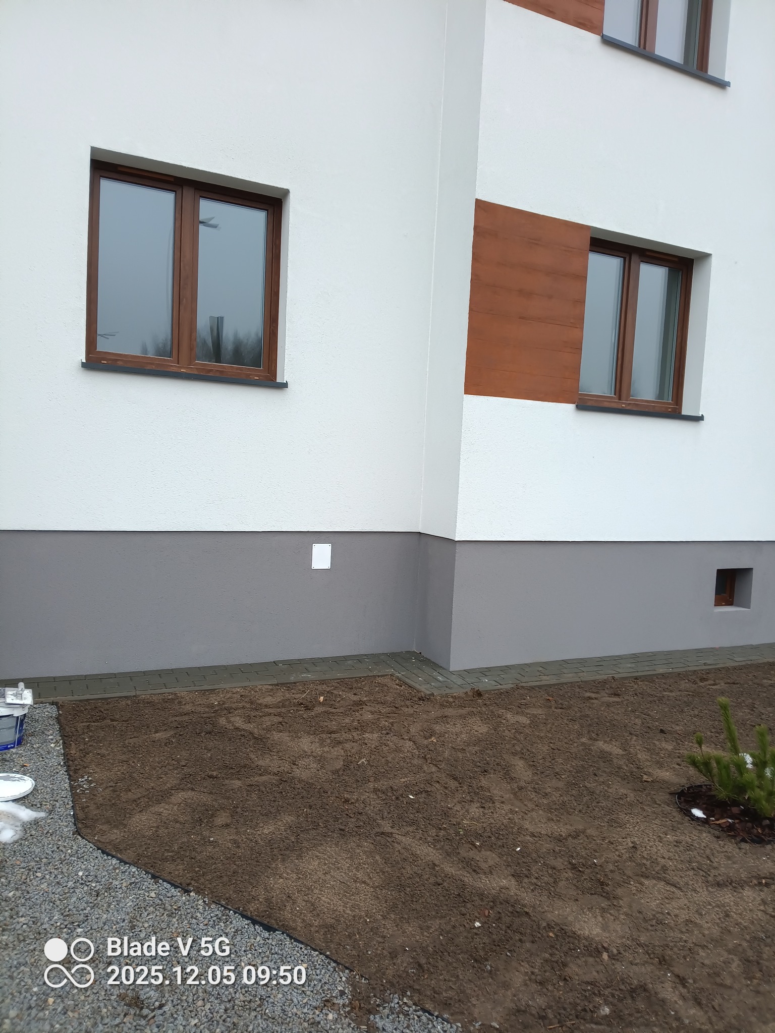 Elewacja domu po malowaniu: biała ściana z brązowymi oknami i szarym cokołem. Świeżo przygotowany teren pod trawnik. Widoczne ślady farby.