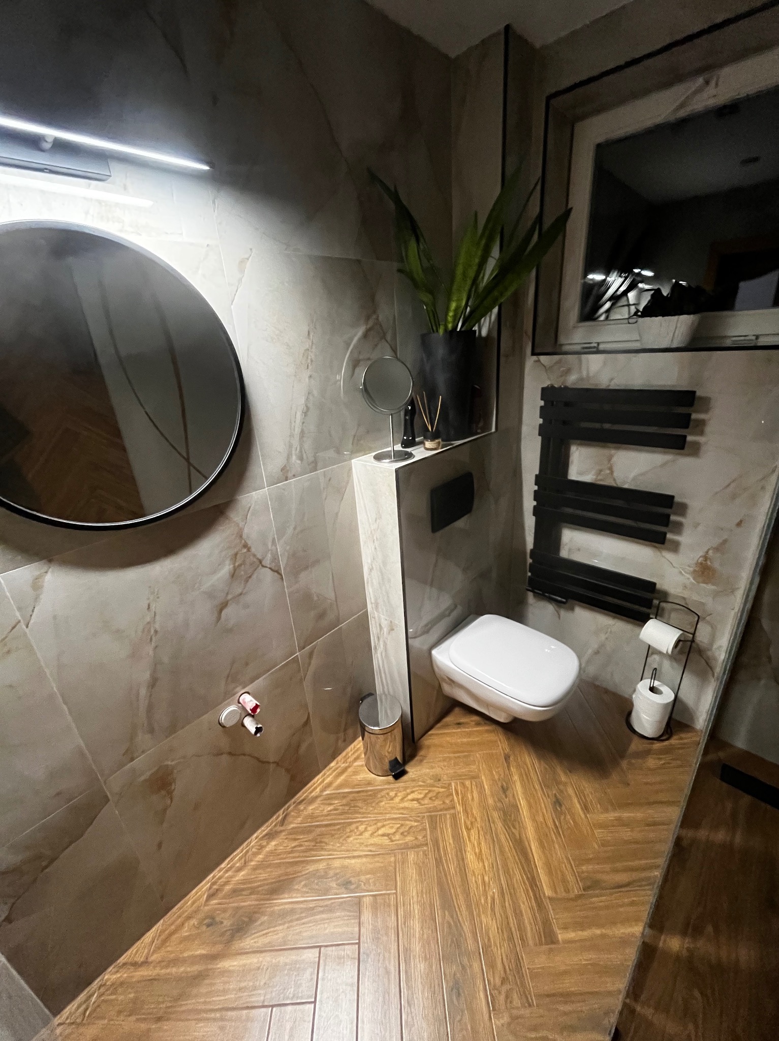 Stylowa łazienka z beżowymi płytkami imitującymi marmur, drewnianą podłogą w jodełkę, okrągłym lustrem i grzejnikiem drabinkowym w kształcie litery E.