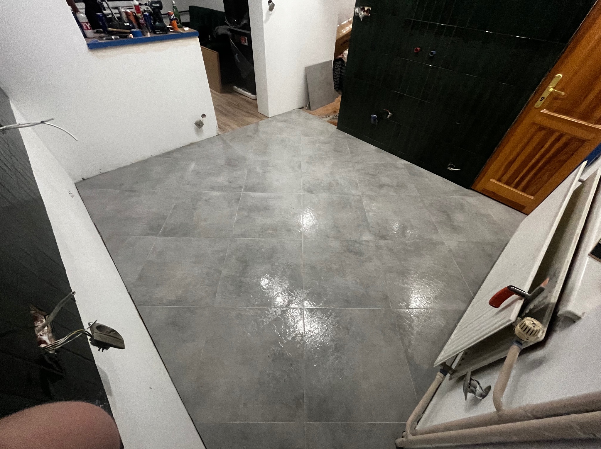 Ułożone szare płytki podłogowe w pomieszczeniu z białymi ścianami i ciemnymi drzwiami. Widoczny fragment grzejnika i instalacji.