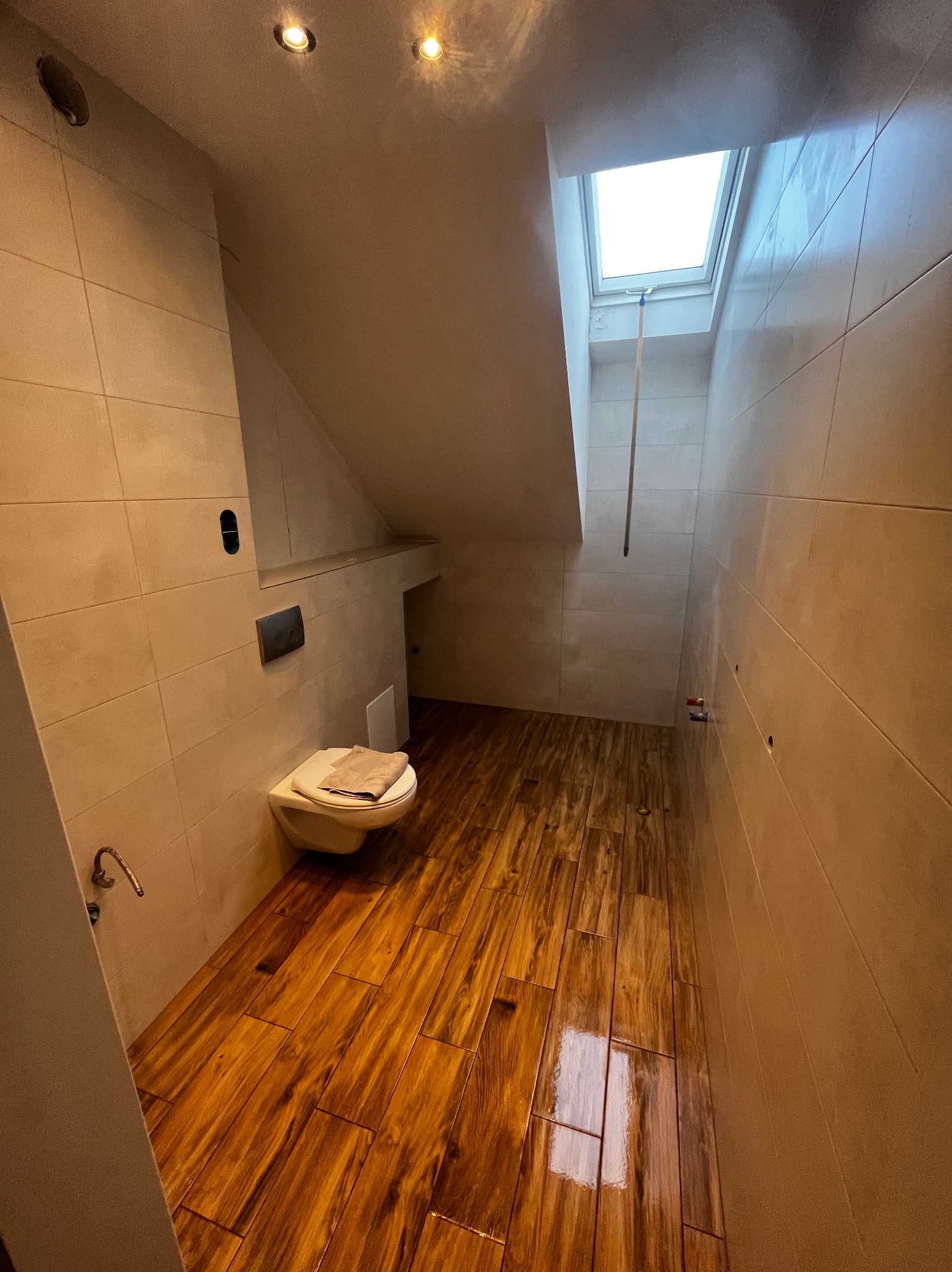 Ukończona łazienka na poddaszu z drewnopodobną podłogą, beżowymi płytkami ściennymi i oknem dachowym. Wisząca toaleta z deską. Oświetlenie punktowe.