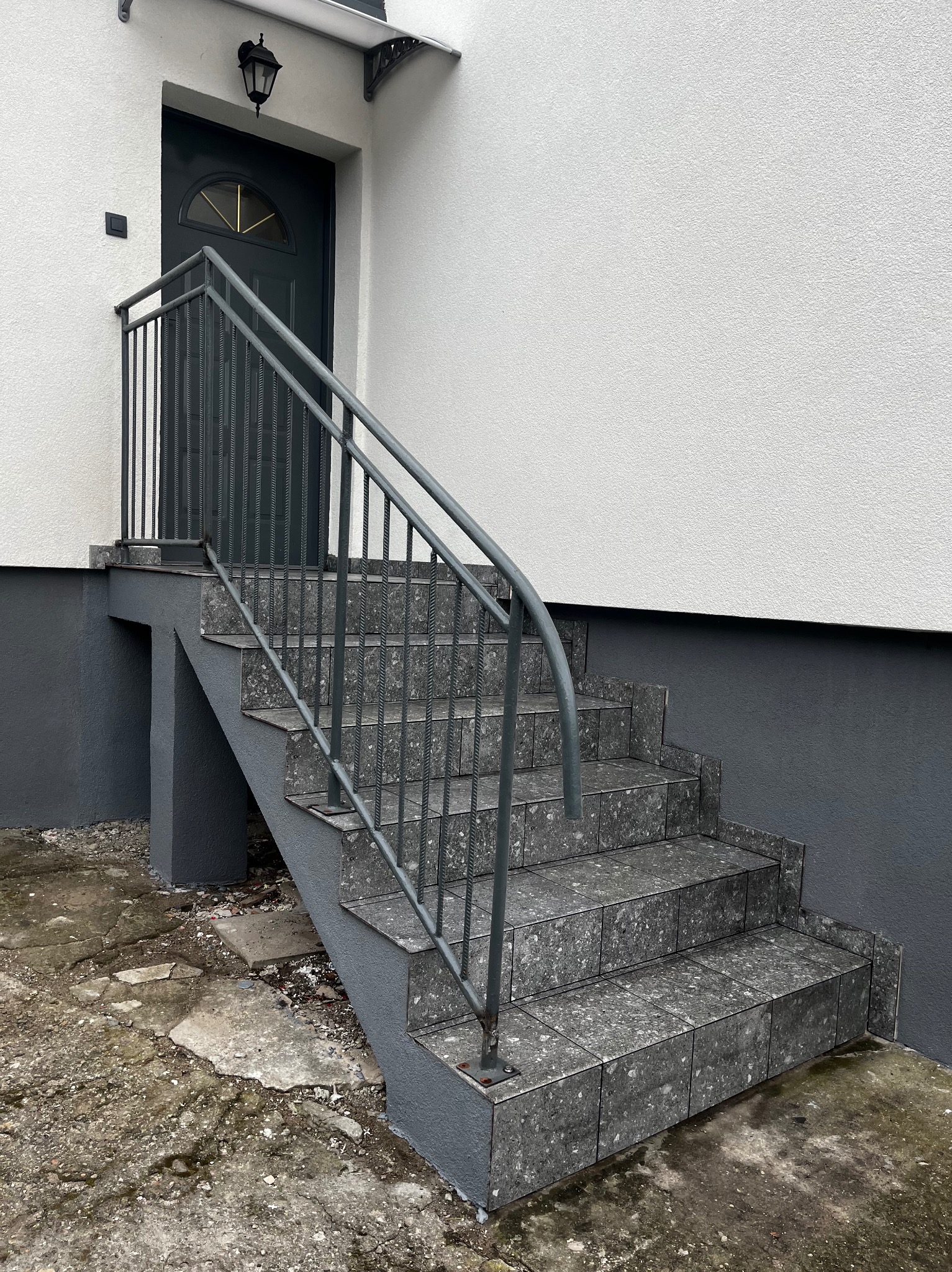 Zewnętrzne schody wejściowe wyłożone szarą, kwadratową terakotą z metalową balustradą. Ściana budynku biała z szarym cokołem. Widok z dołu, lekko z prawej strony.