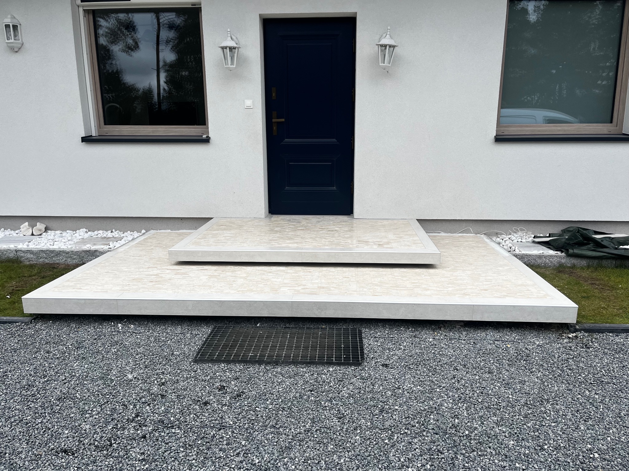 Nowoczesne, dwupoziomowe schody wejściowe z jasnych płytek, prowadzące do ciemnych drzwi wejściowych w białym budynku. Widoczne okna i szare kamienie na podłożu.