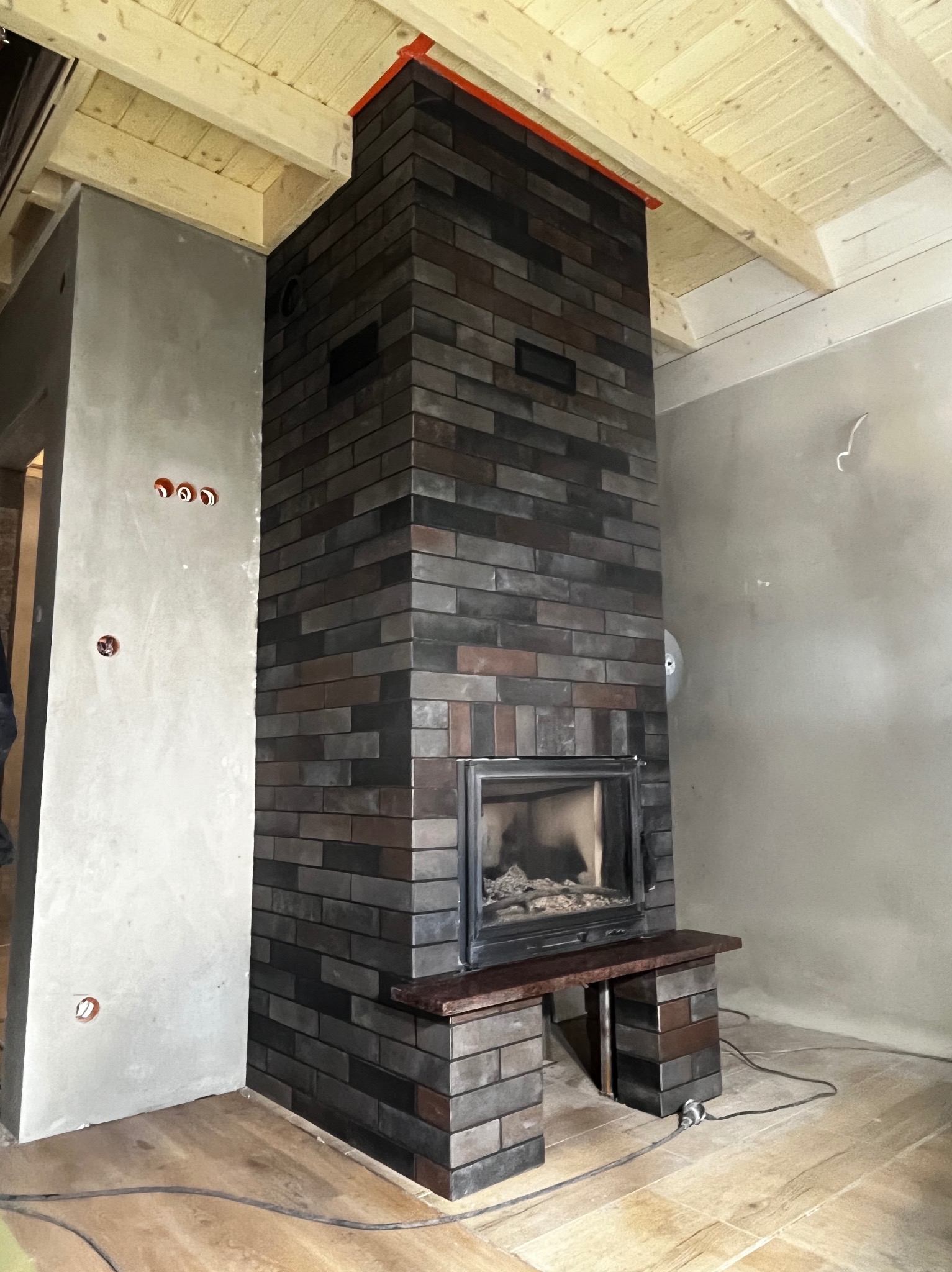 Murowany kominek z ciemnej cegły w surowym wnętrzu. Widoczne niedokończone ściany i drewniany sufit. Otwory na gniazdka elektryczne na ścianie.