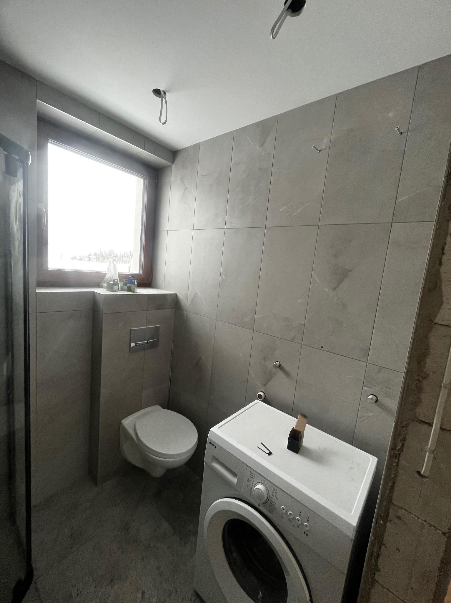 Jasna łazienka w trakcie wykańczania: szare płytki, podwieszana toaleta, pralka z narzędziami na górze. Widoczne niedokończone elementy instalacji.