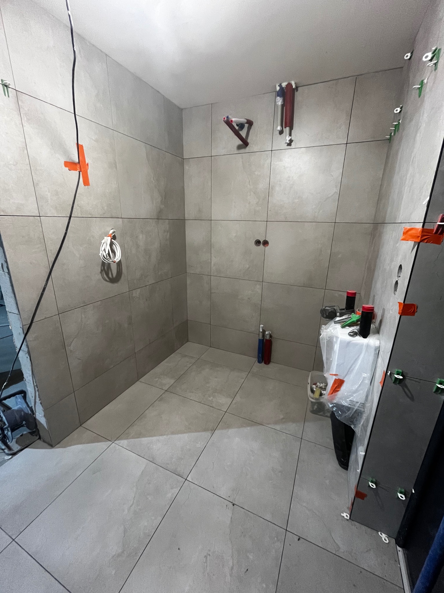 Szara łazienka w trakcie wykańczania: ułożone płytki, widoczne narzędzia glazurnicze i elementy instalacji. Pomarańczowe krzyżyki dystansowe na ścianach.