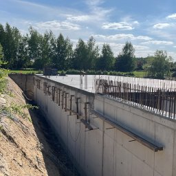 Ivan Pokurbanych - Fundamenty budynku w trakcie budowy, widoczne zbrojenia i betonowe ściany szalunkowe, ziemia, drzewa w tle, słoneczny dzień.