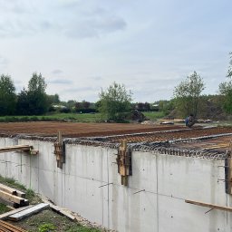 Ivan Pokurbanych - Zbrojenie płyty fundamentowej z betonu, widoczne drewniane elementy szalunku i dwóch pracowników na budowie. W tle zieleń drzew i pochmurne niebo.