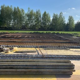 Ivan Pokurbanych - Zbrojenie fundamentów na żółtej płycie szalunkowej, widok z góry. W tle wykop i zieleń drzew. Prace przygotowawcze do wylania betonu.