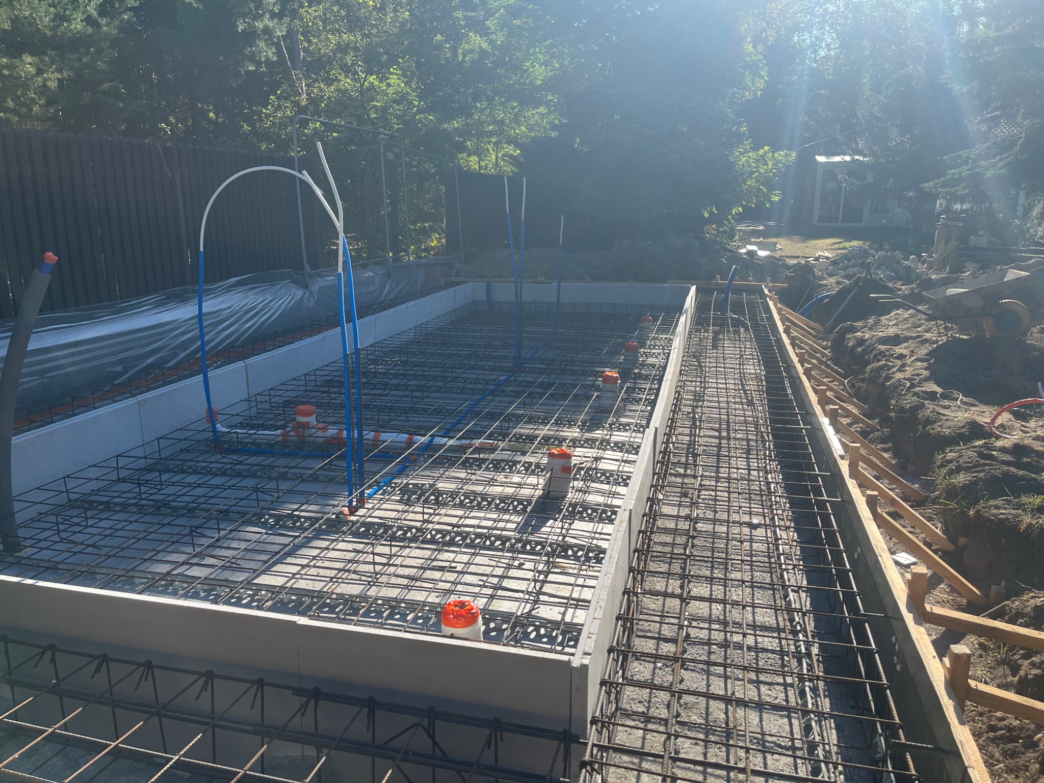 Zbrojenie fundamentów pod budowę domu jednorodzinnego. Widoczne szalunki, siatka zbrojeniowa i wystające rury instalacyjne. Słoneczny dzień.