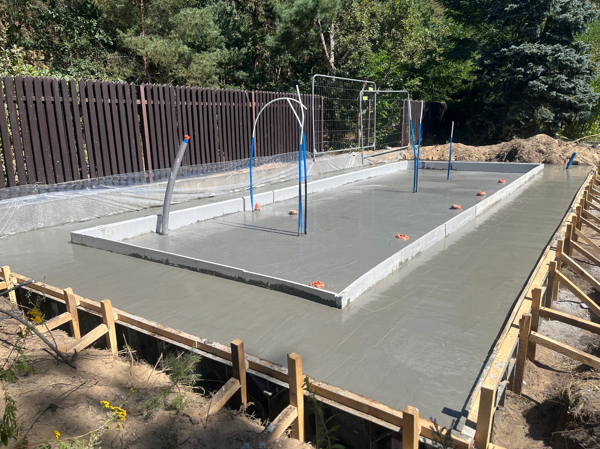Zalany fundament pod budowę domu jednorodzinnego, widoczne szalunki i wystające rury instalacyjne. Świeży beton w ramie fundamentowej, ogrodzenie w tle.