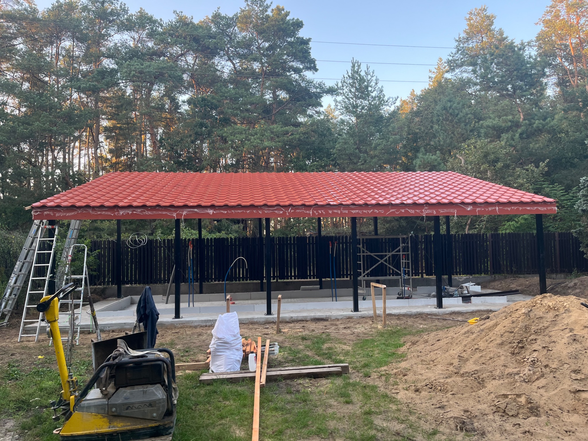 Budowa zadaszenia w Warszawie: widok na konstrukcję z czerwoną dachówką, czarne słupy i częściowo wylane fundamenty. Na pierwszym planie zagęszczarka.