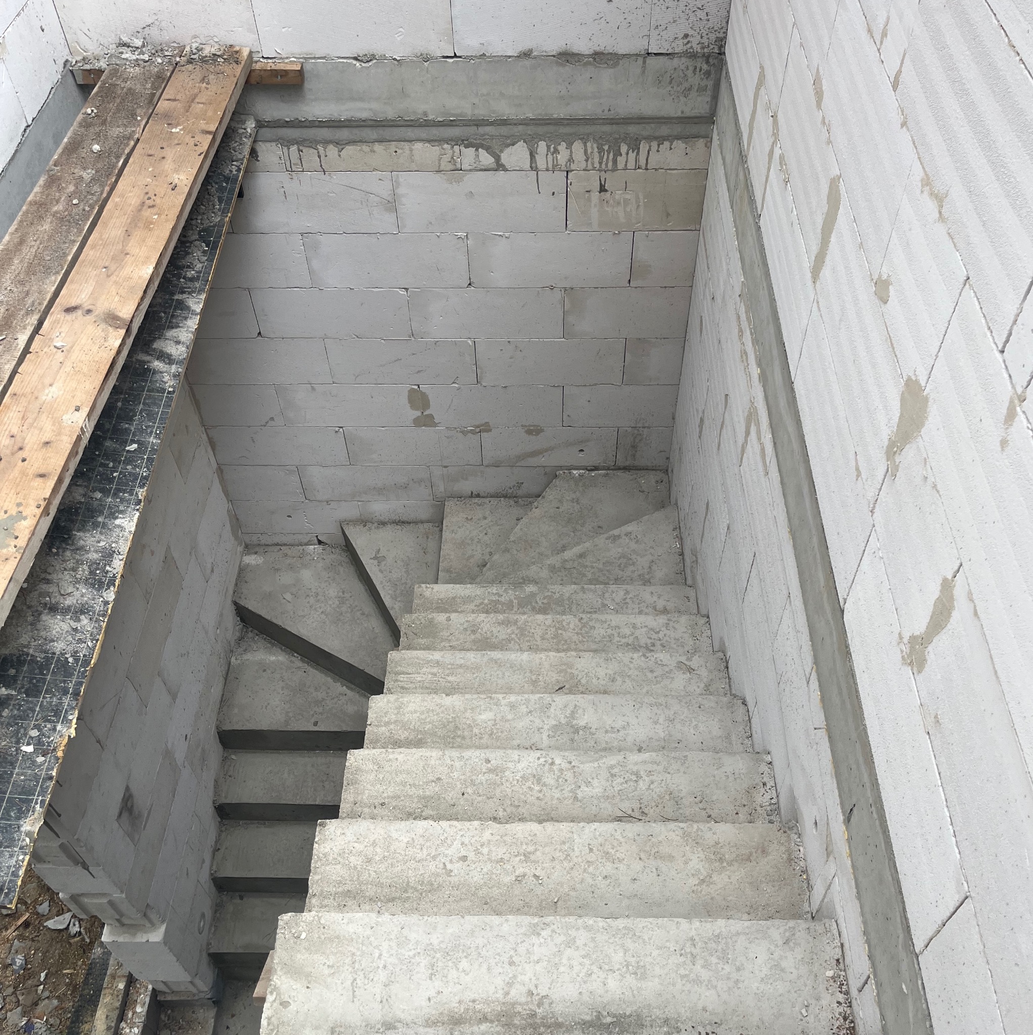 Betonowe schody w surowym stanie, prowadzące w dół, otoczone ścianami z bloczków betonowych. Widoczne ślady zaprawy i drewniane elementy szalunku.
