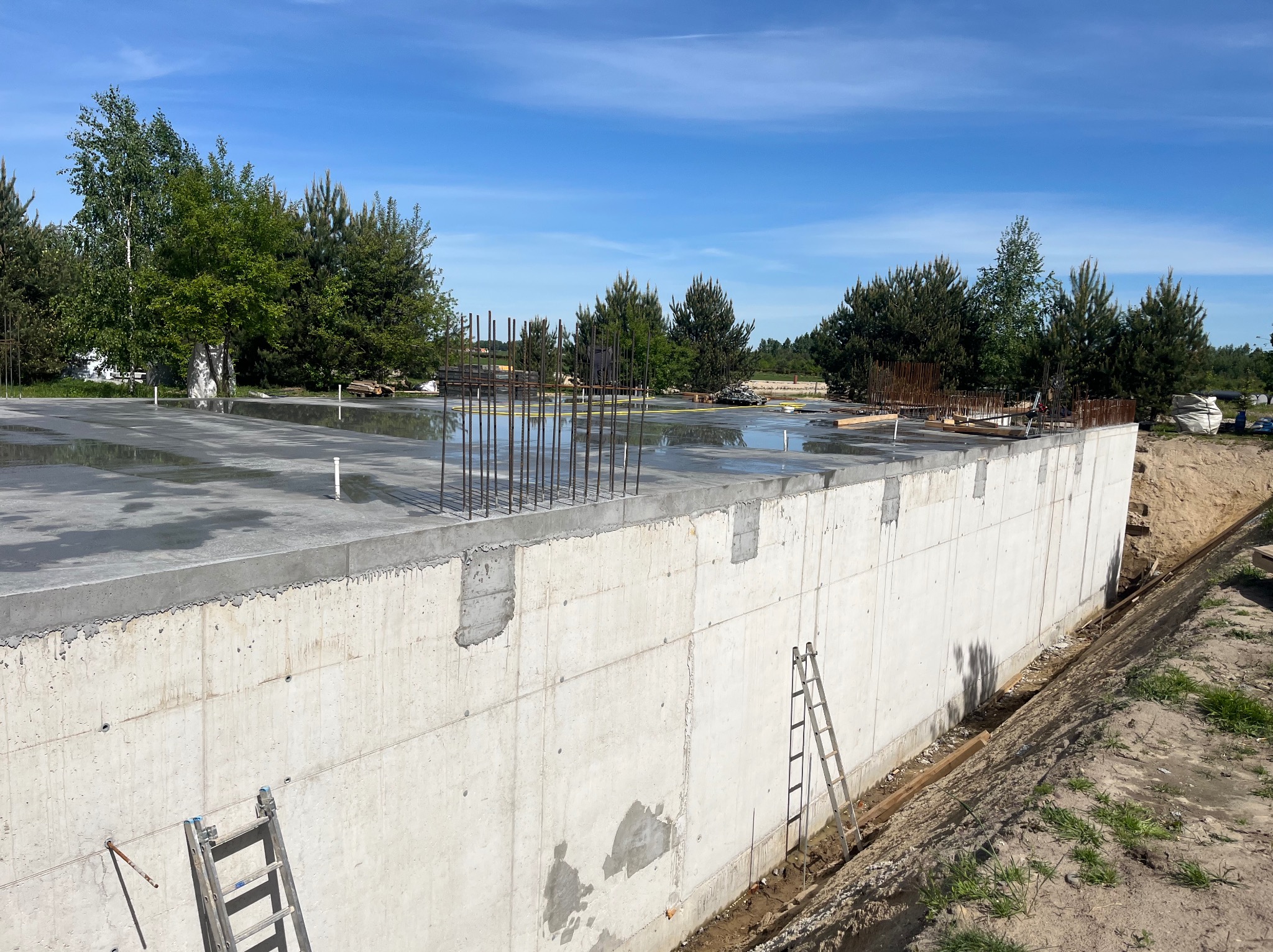 Wylana płyta fundamentowa z wystającymi prętami zbrojeniowymi, widoczna ściana fundamentowa z drabinkami opartymi o nią. W tle zieleń drzew i błękitne niebo.
