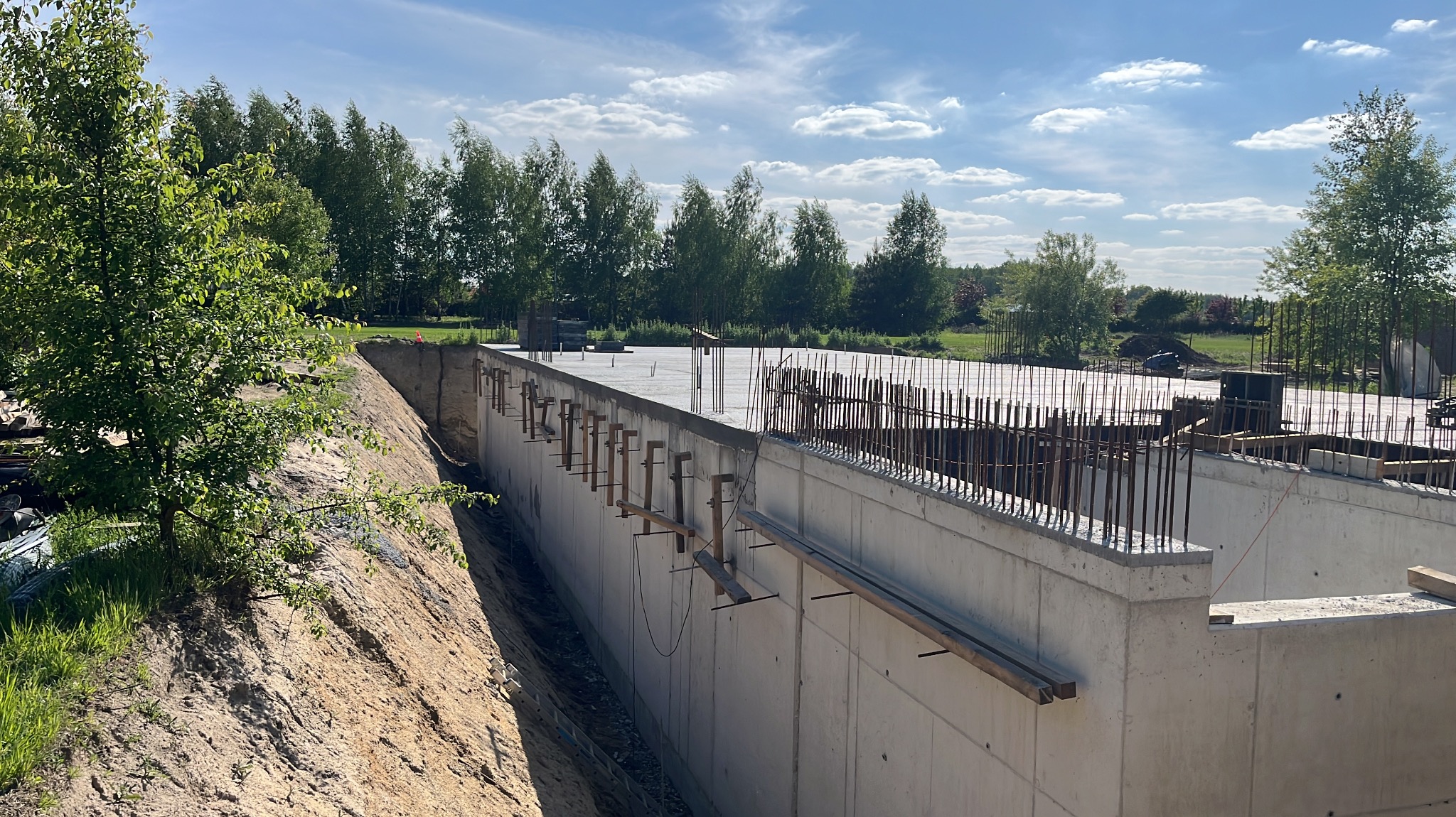 Fundamenty budynku w trakcie budowy, widoczne zbrojenia i betonowe ściany szalunkowe, ziemia, drzewa w tle, słoneczny dzień.