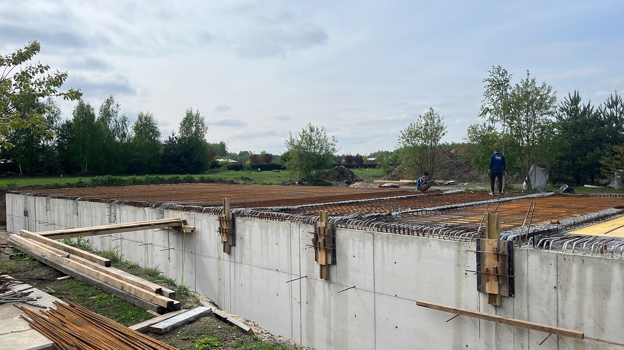 Zbrojenie płyty fundamentowej z betonu, widoczne drewniane elementy szalunku i dwóch pracowników na budowie. W tle zieleń drzew i pochmurne niebo.